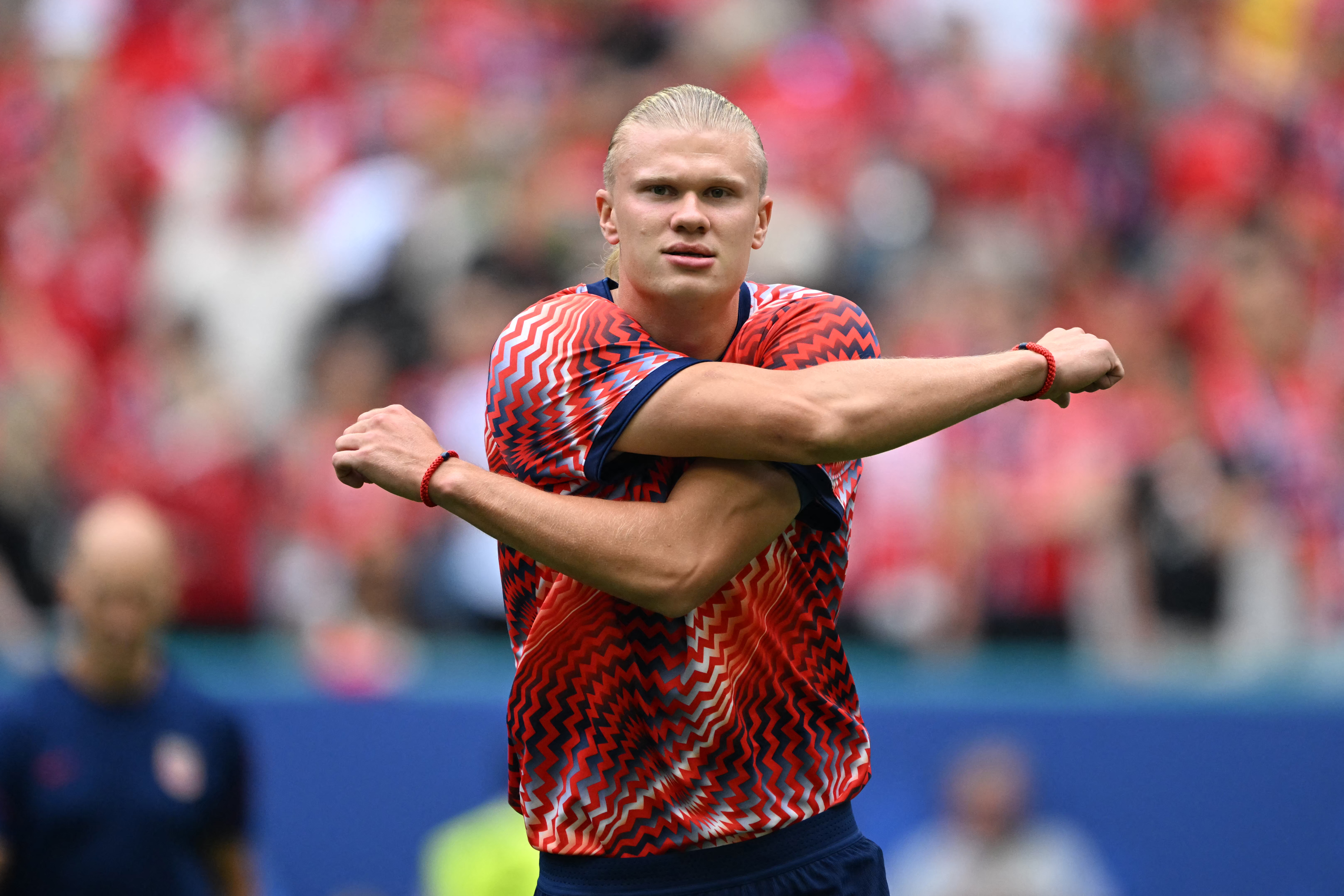 Erling Haaland in a red patterned training top stretches his arms across his chest on the pitch before a match.
