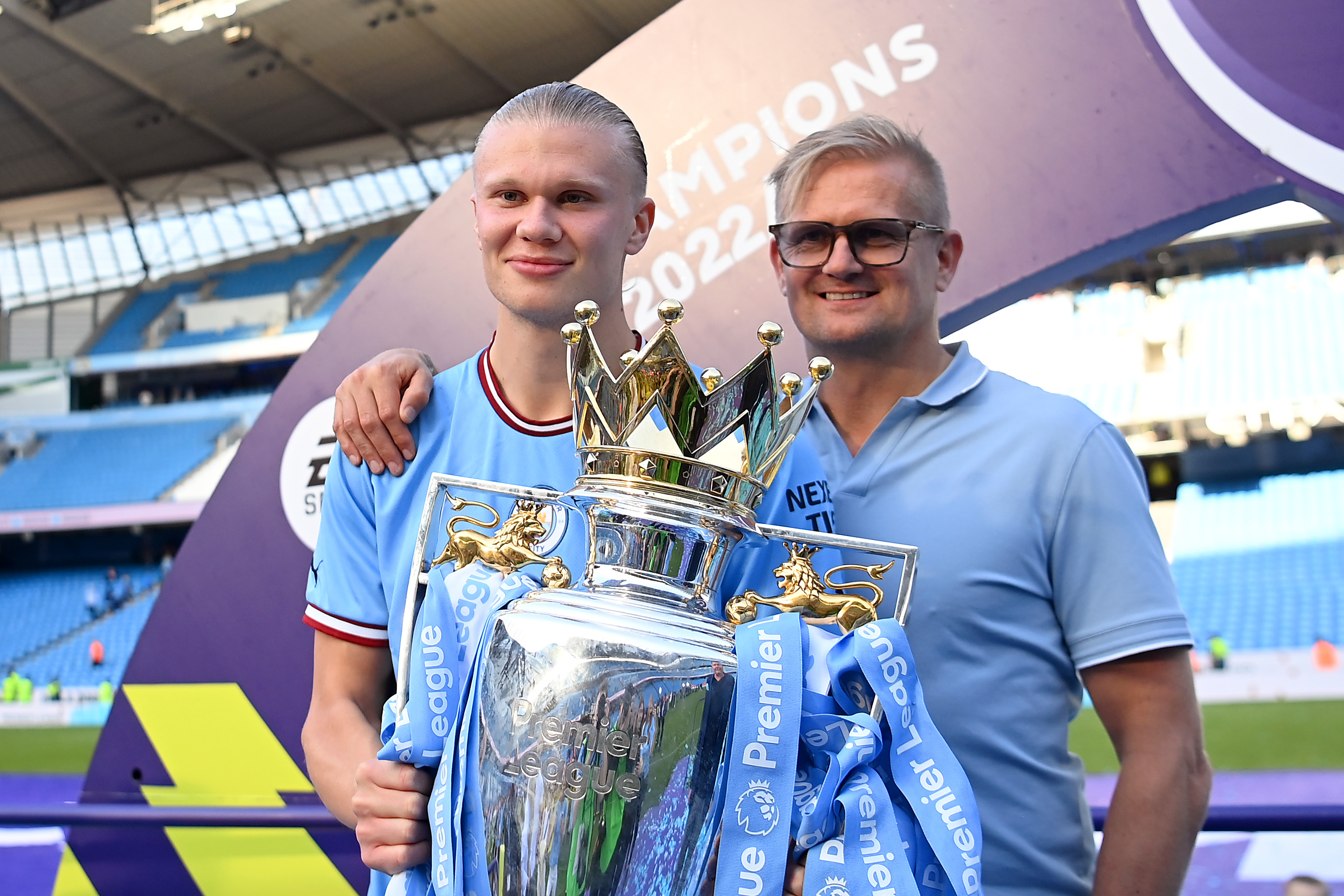 Erling Haaland in a Manchester City kit holds the Premier League Trophy alongside his father Alf-Inge Haaland, both smiling in front of a Champions 2022 banner at Etihad Stadium.