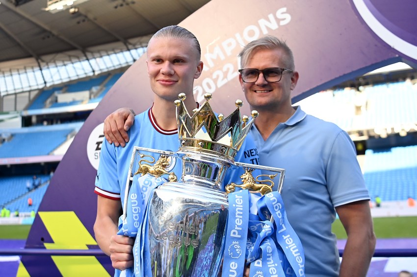 Erling Haaland in a Manchester City kit holds the Premier League Trophy alongside his father Alf-Inge Haaland, both smiling in front of a Champions 2022 banner at Etihad Stadium.