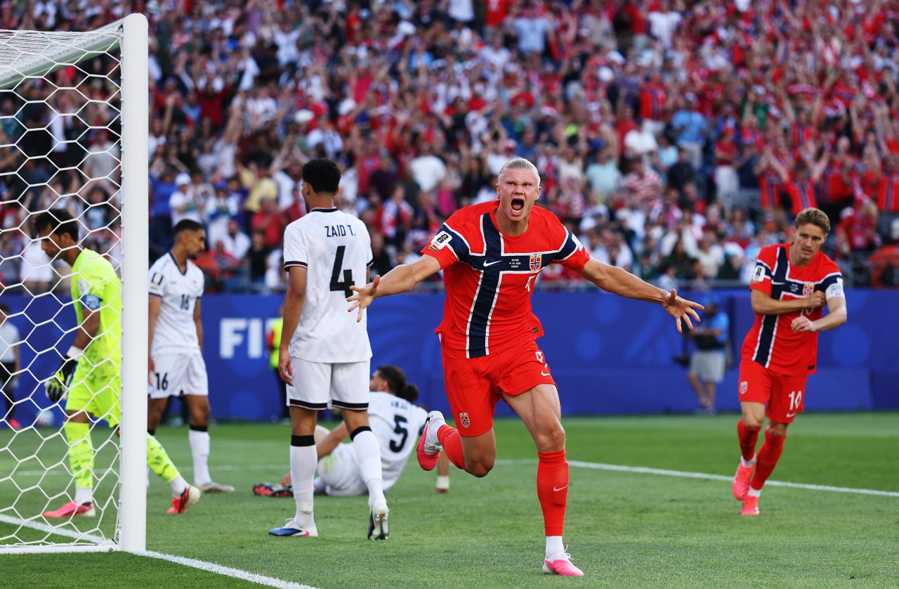 Erling Haaland in a red Norway kit screaming in celebration near the goal post, arms spread wide, with a packed stadium crowd behind him.