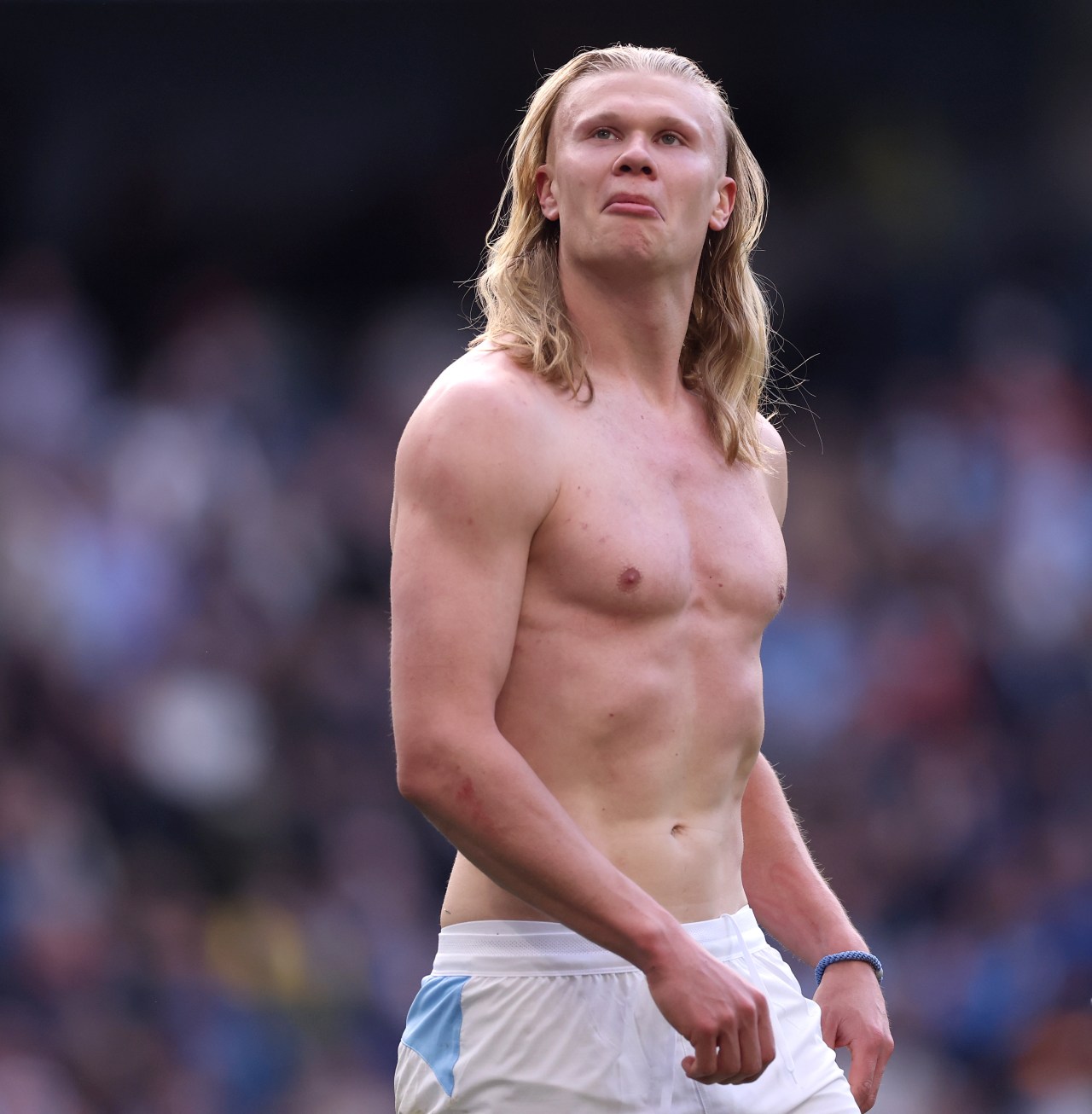 Shirtless Erling Haaland with long blond hair looks upward on a football pitch, wearing white Manchester City shorts, crowd blurred behind him.