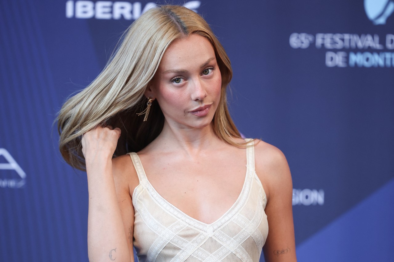 Blonde woman in a cream spaghetti-strap mesh dress brushing hair from her face in front of a blue Monte-Carlo Television Festival banner.