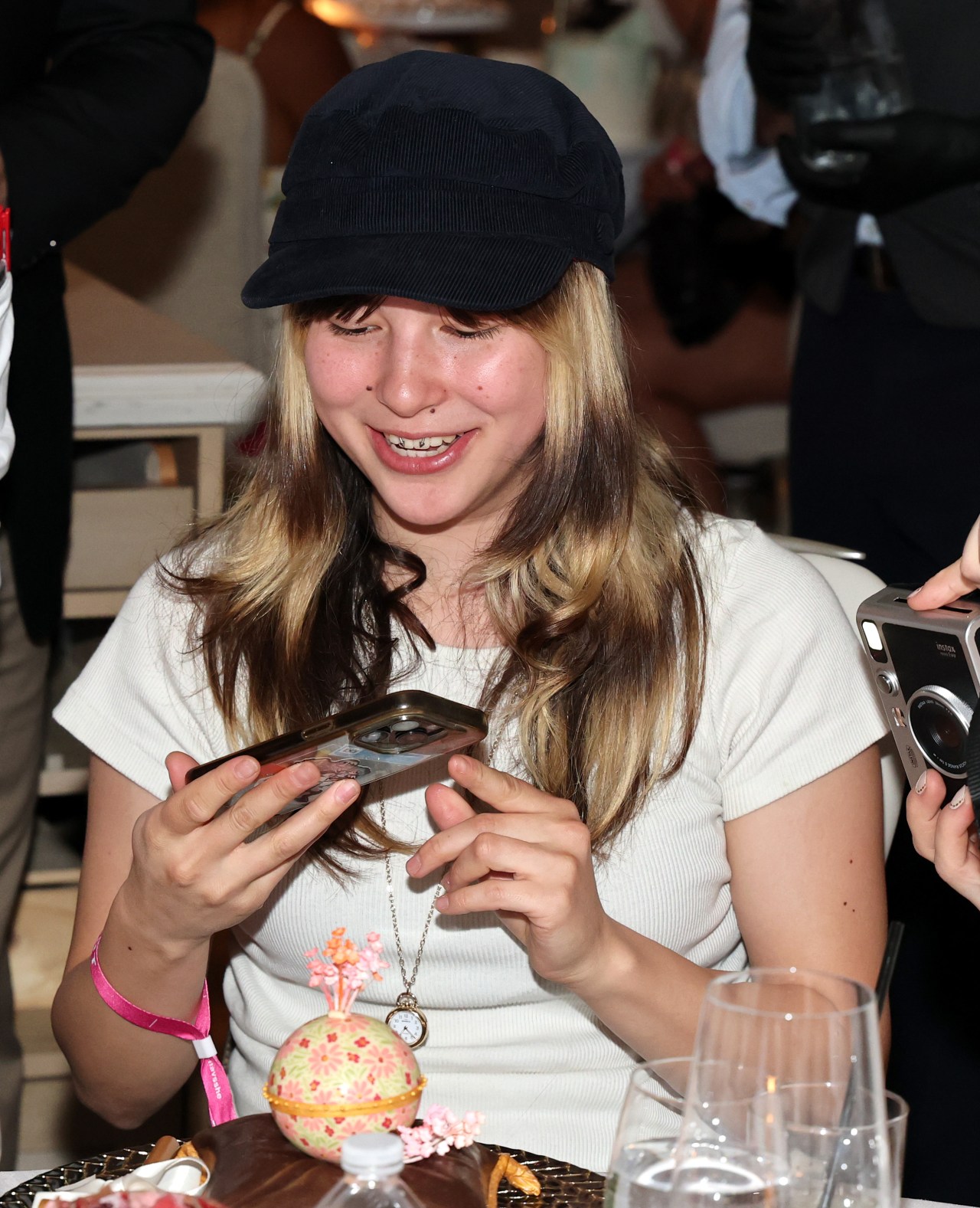 Young woman in a navy newsboy cap smiling while photographing a floral chocolate sculpture on a dinner table.