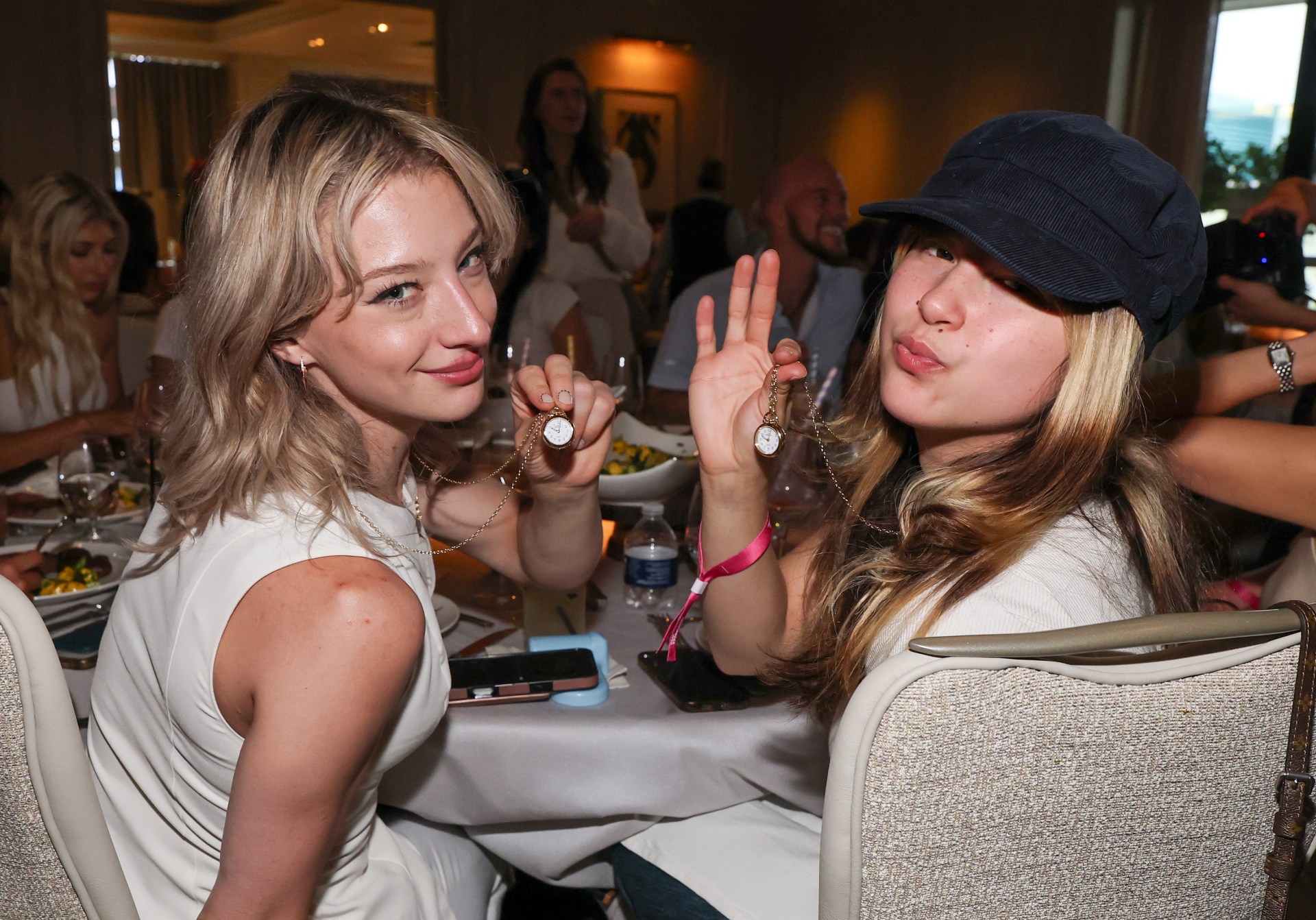 Two young women seated at a dinner table holding up small clock necklaces on chains, one in a white top and one in a cap making a kissy face.