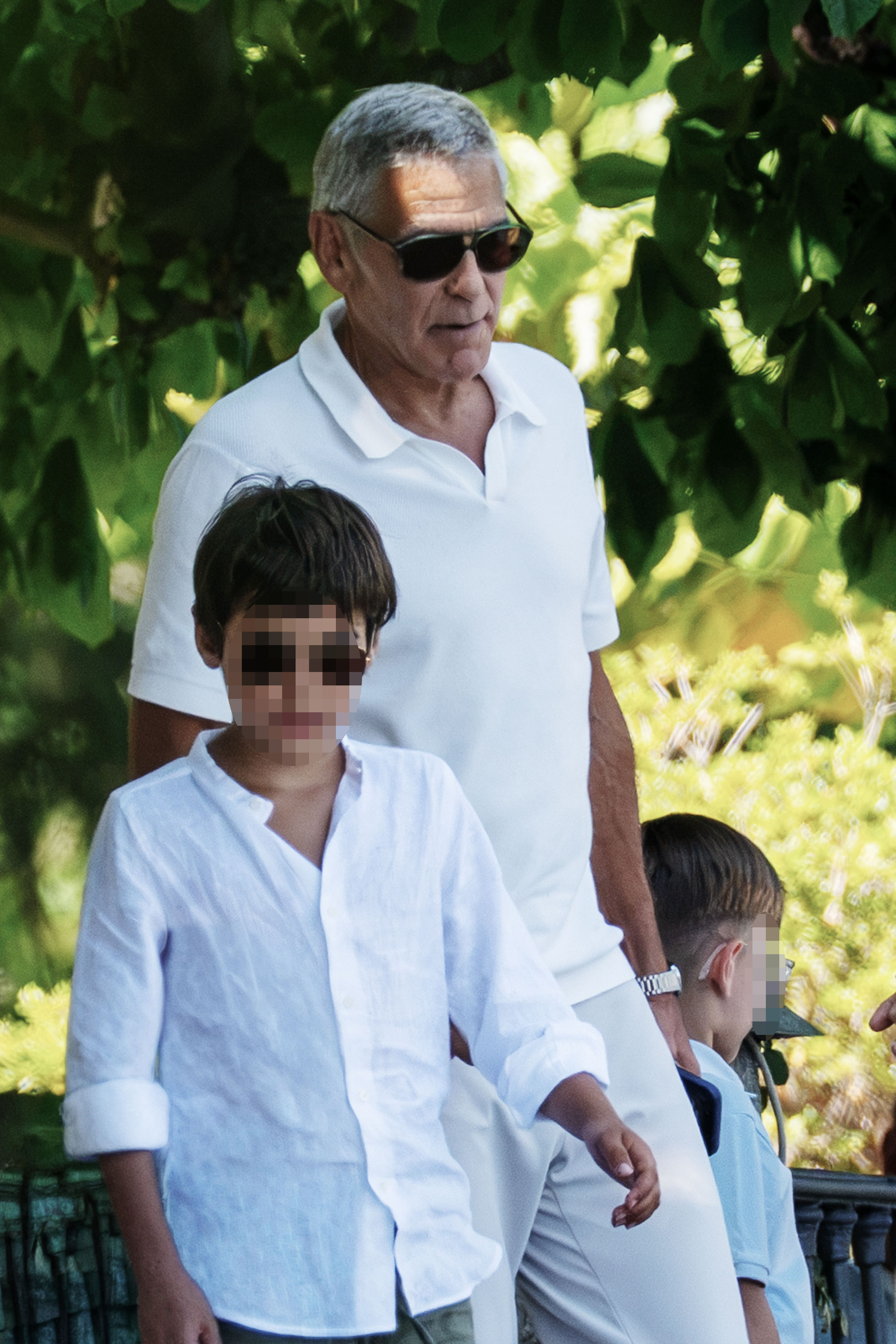 An older man with silver hair and sunglasses in a white polo shirt walks with two young children, both with pixelated faces, outdoors near lush greenery.