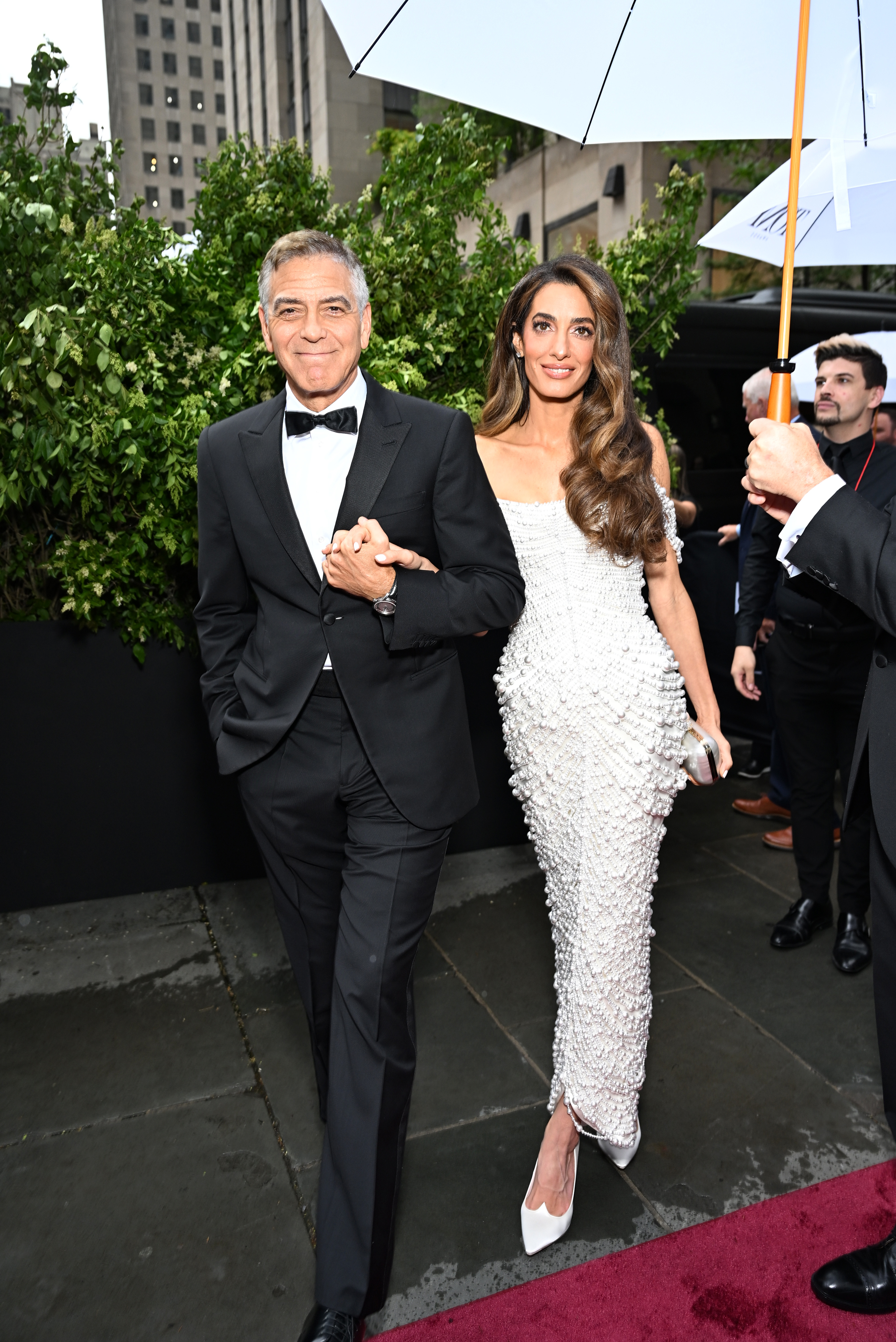 George Clooney in a black tuxedo and Amal Clooney in a white beaded gown walking the red carpet at the Tony Awards in New York City.