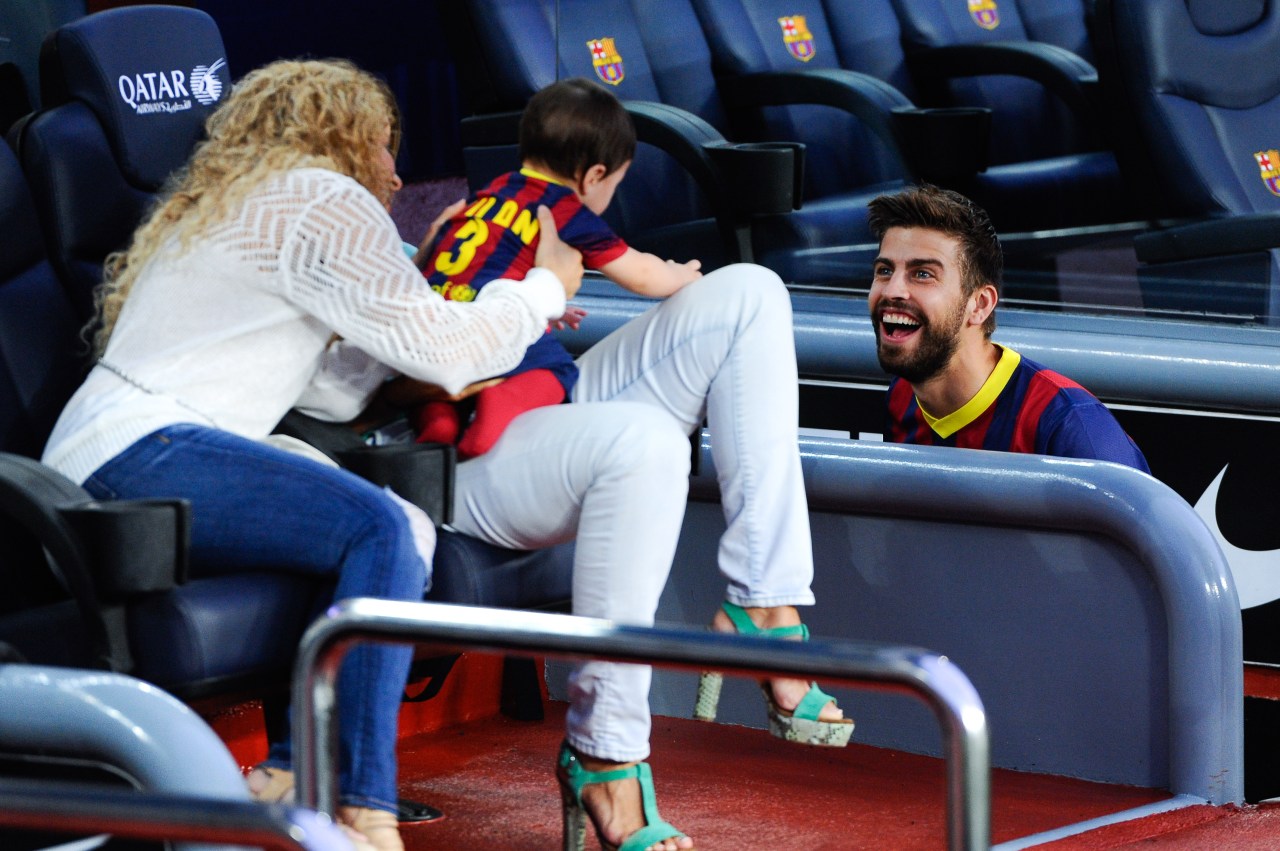 Gerard Piqué laughing while seated at Camp Nou as Shakira holds baby Milan in a FC Barcelona jersey in the stands beside him.