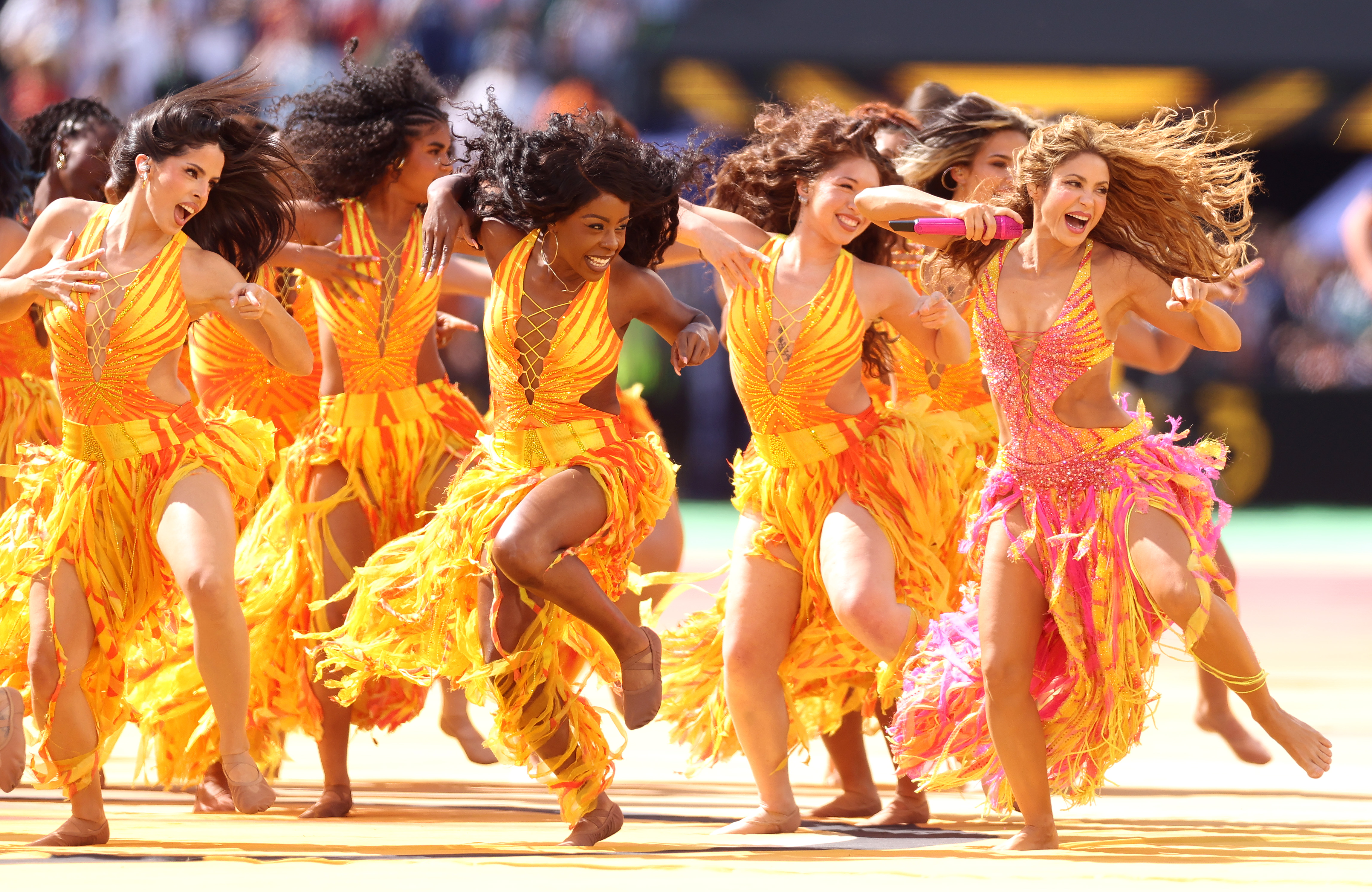 Shakira in a pink and gold sequined costume holds a microphone while dancing alongside performers in orange feathered outfits at a stadium halftime show.