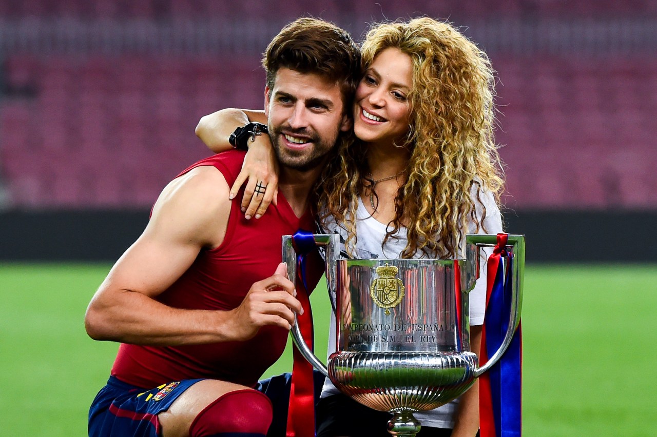 Gerard Piqué in a red Barcelona kit and Shakira with curly hair crouching together beside the Copa del Rey trophy on a football pitch.