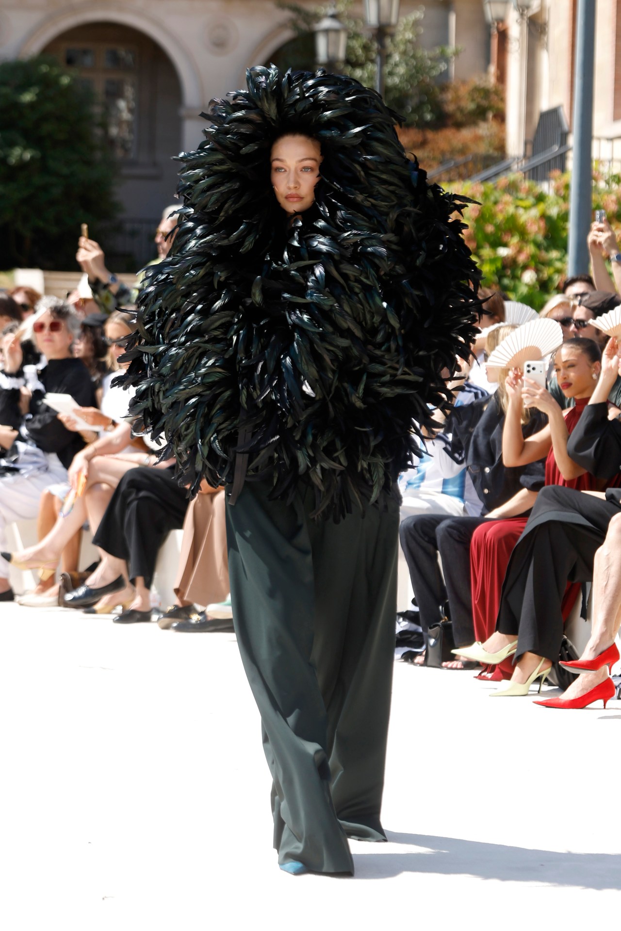 Model walking a fashion runway wearing a dramatic oversized black feather cape that frames her face and torso, paired with a dark green floor-length gown, before a seated crowd.