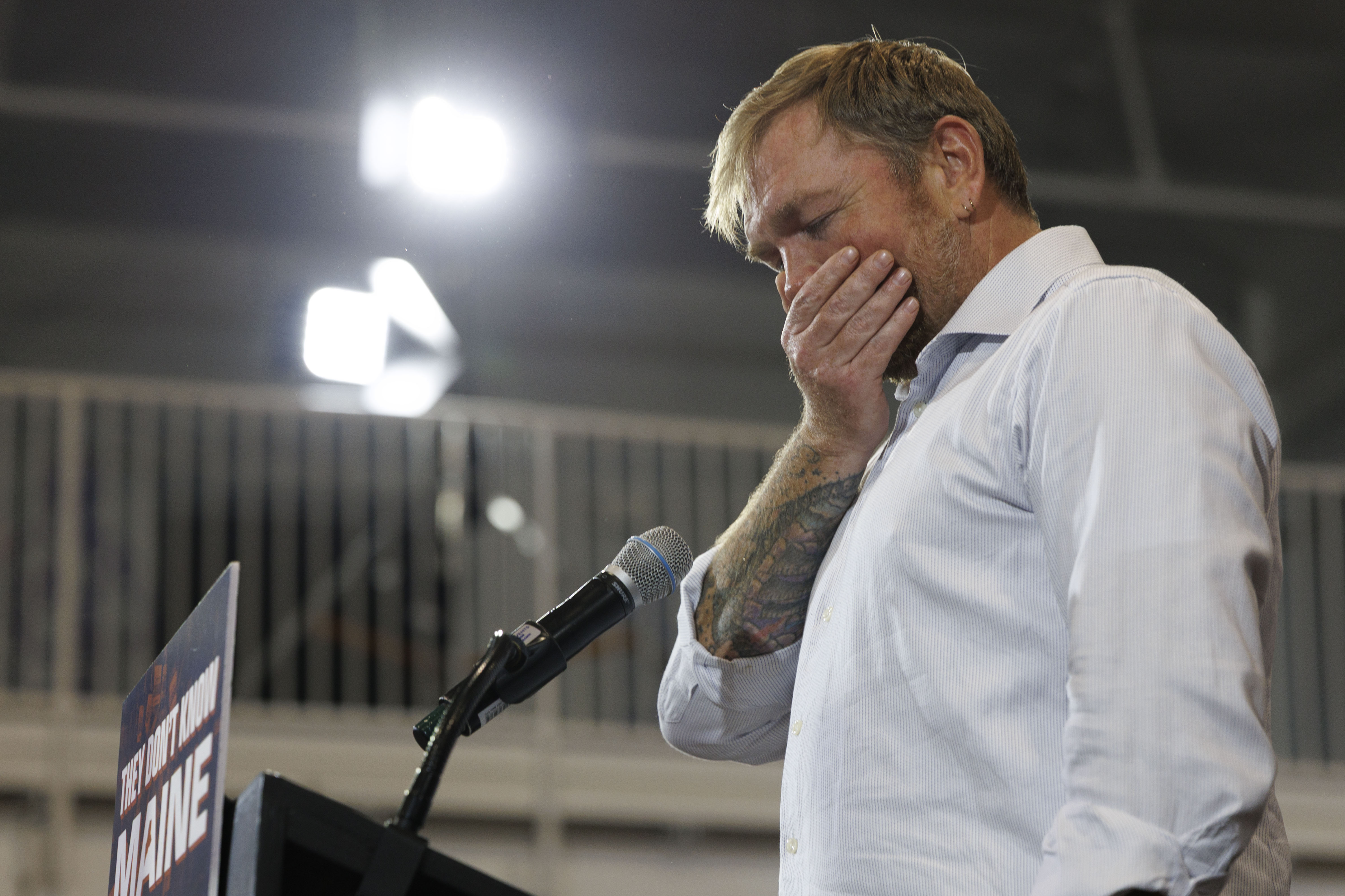Man in white shirt with tattooed arm covers his face with one hand while standing at a microphone podium with a 'They Don't Know Maine' sign.