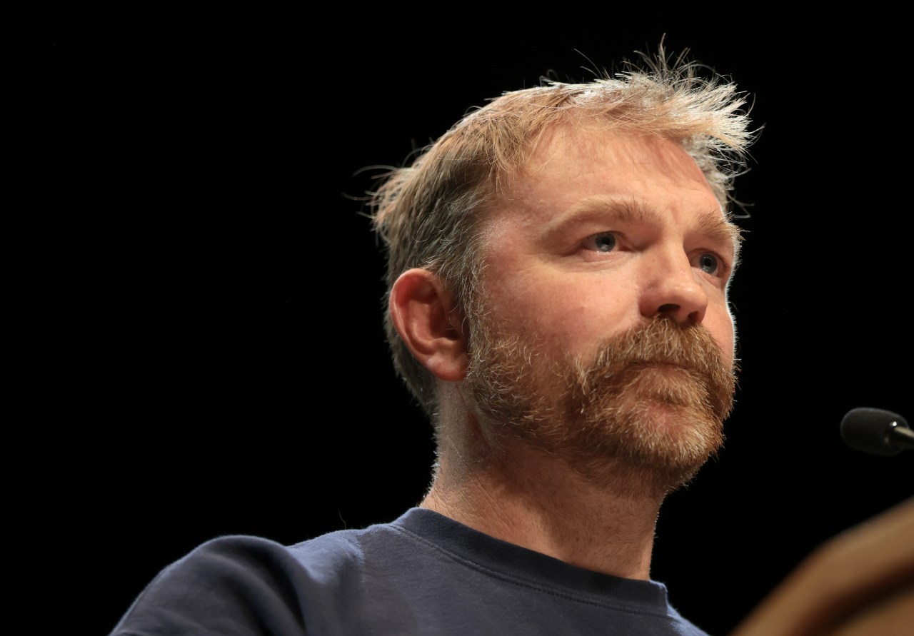 Bearded man in a blue t-shirt gazes upward from a podium against a dark background, with a microphone visible at the right edge.