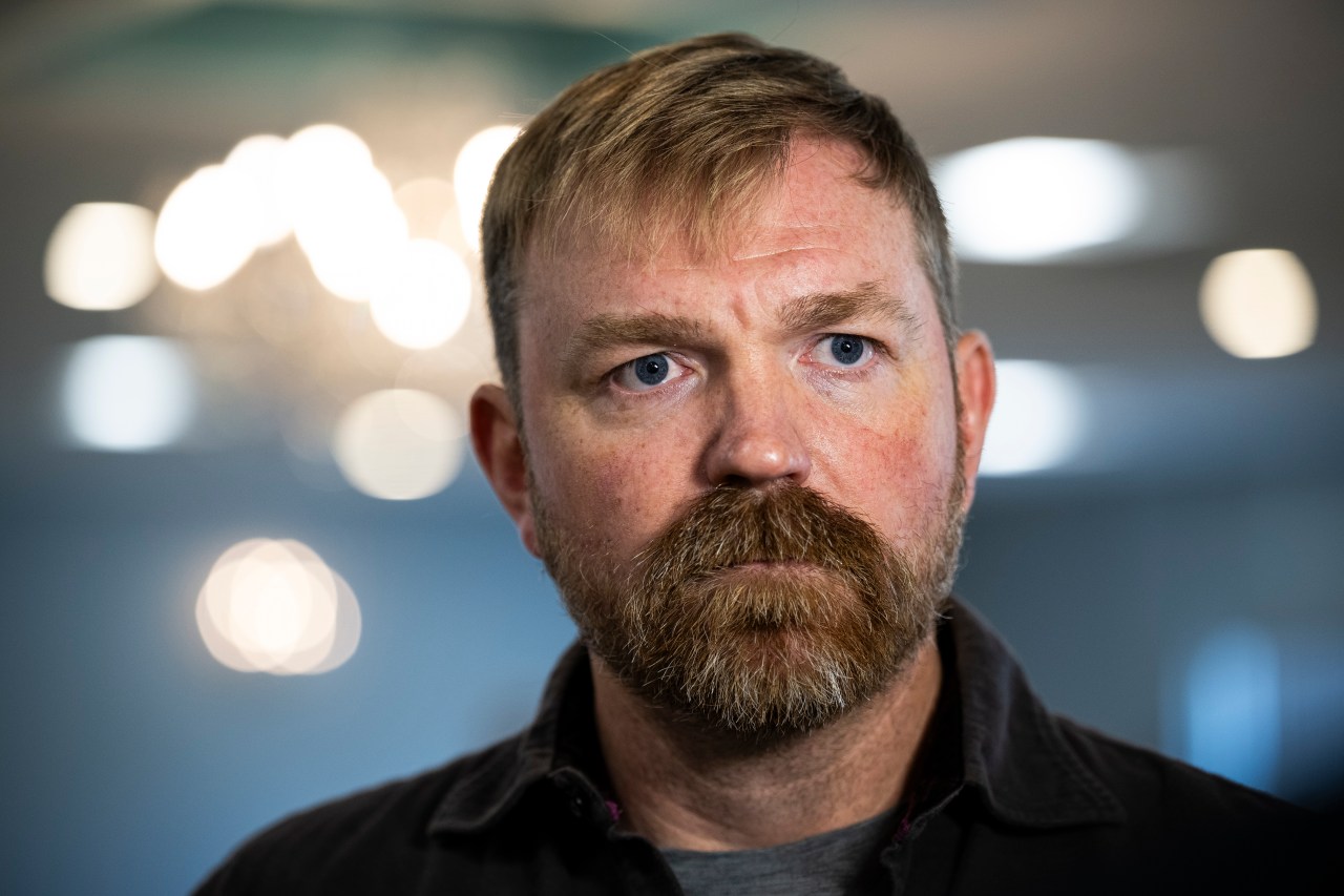 Close-up portrait of a man with blue eyes and a reddish goatee in a dark jacket, with bokeh lights in the background.