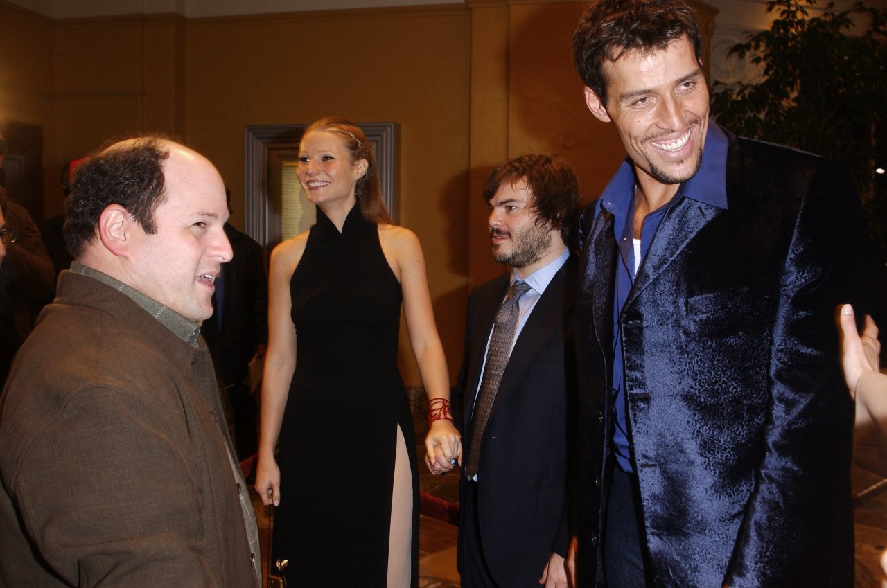 Four people posing at a movie premiere: a bald man in brown coat, a tall blonde woman in black dress, a shorter bearded man in suit, and a tall man in blue blazer.
