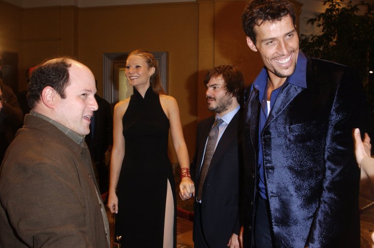 Four people posing at a movie premiere: a bald man in brown coat, a tall blonde woman in black dress, a shorter bearded man in suit, and a tall man in blue blazer.