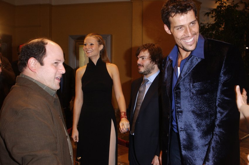 Four people posing at a movie premiere: a bald man in brown coat, a tall blonde woman in black dress, a shorter bearded man in suit, and a tall man in blue blazer.