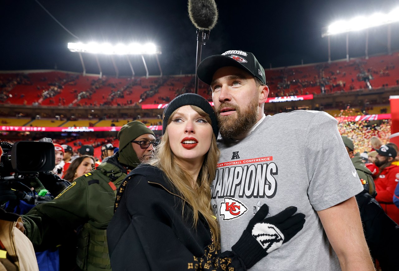 Taylor Swift in a black beanie and jacket leans against Travis Kelce, who wears a Kansas City Chiefs AFC Champions t-shirt, on the Arrowhead Stadium field at night.