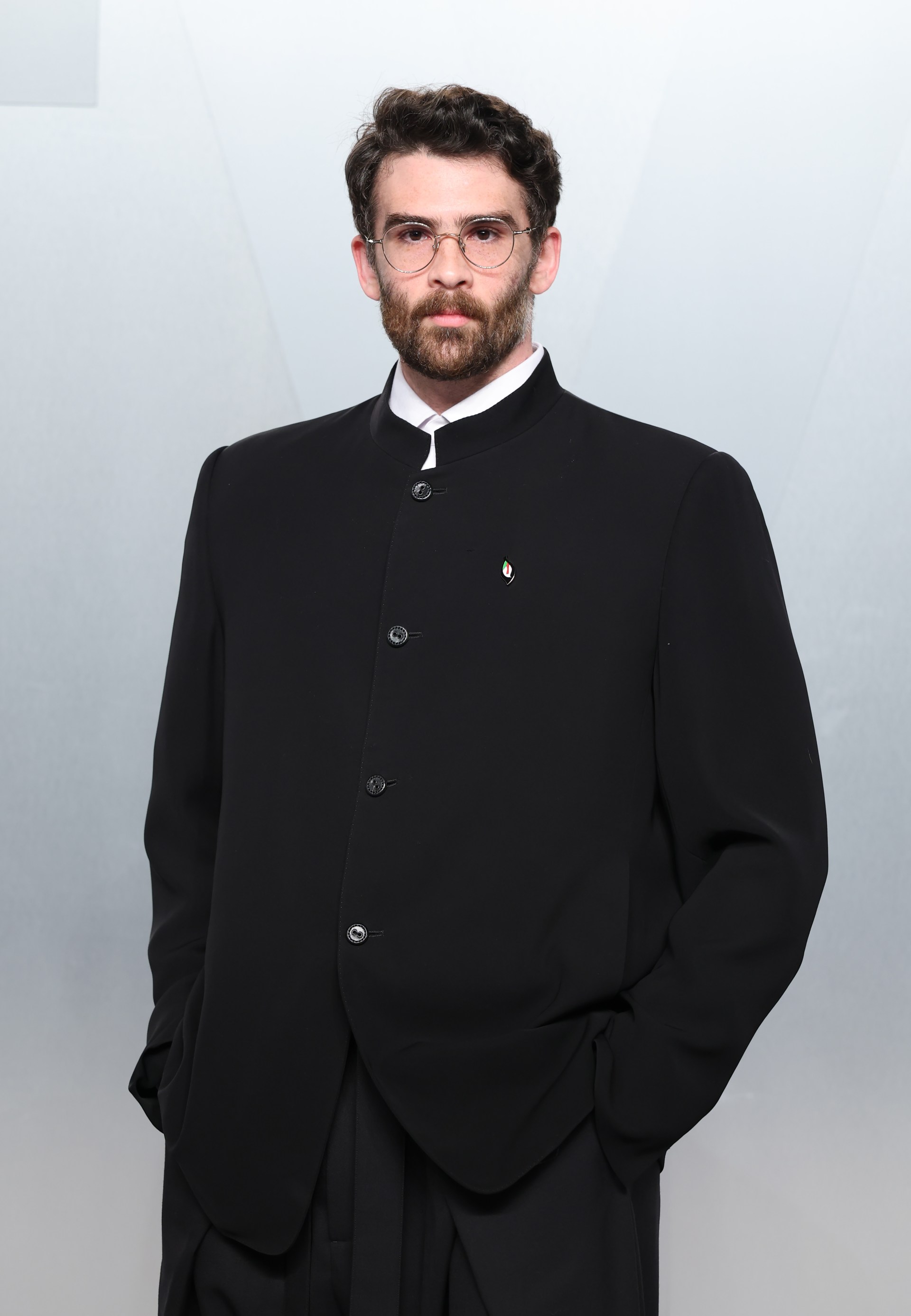 Man with curly brown hair, beard, and round glasses wearing a black mandarin-collar jacket with a small lapel pin, standing against a gray backdrop.
