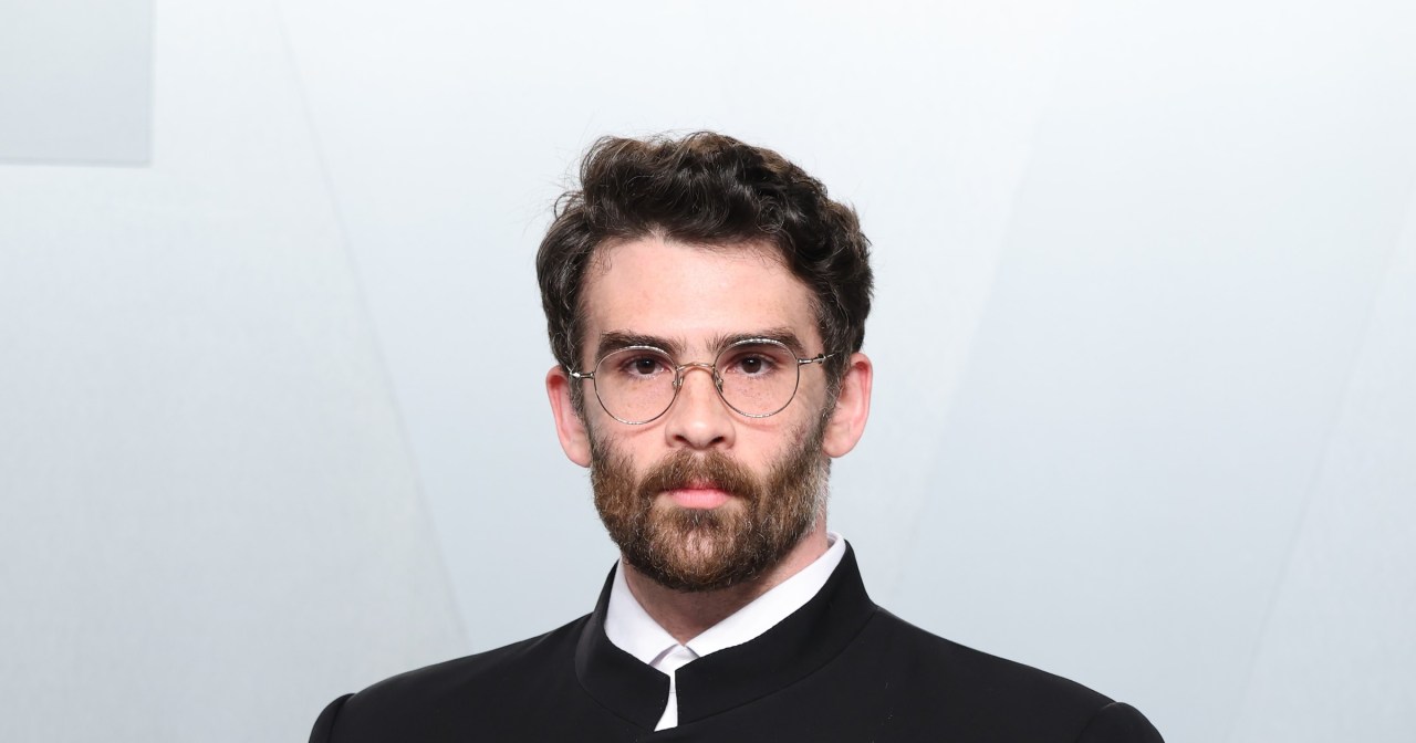 Hasan Piker wearing a black mandarin-collar jacket with a small lapel pin, hands in pockets, at the 2026 Vanity Fair Oscar Party in Los Angeles.