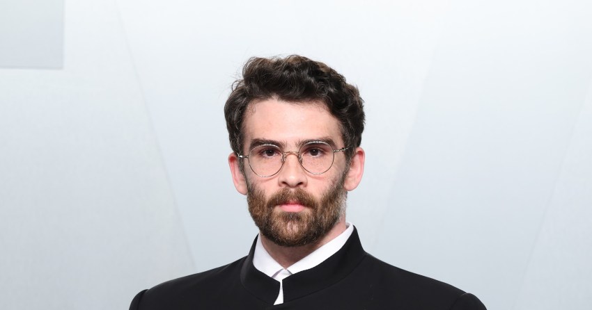 Hasan Piker wearing a black mandarin-collar jacket with a small lapel pin, hands in pockets, at the 2026 Vanity Fair Oscar Party in Los Angeles.