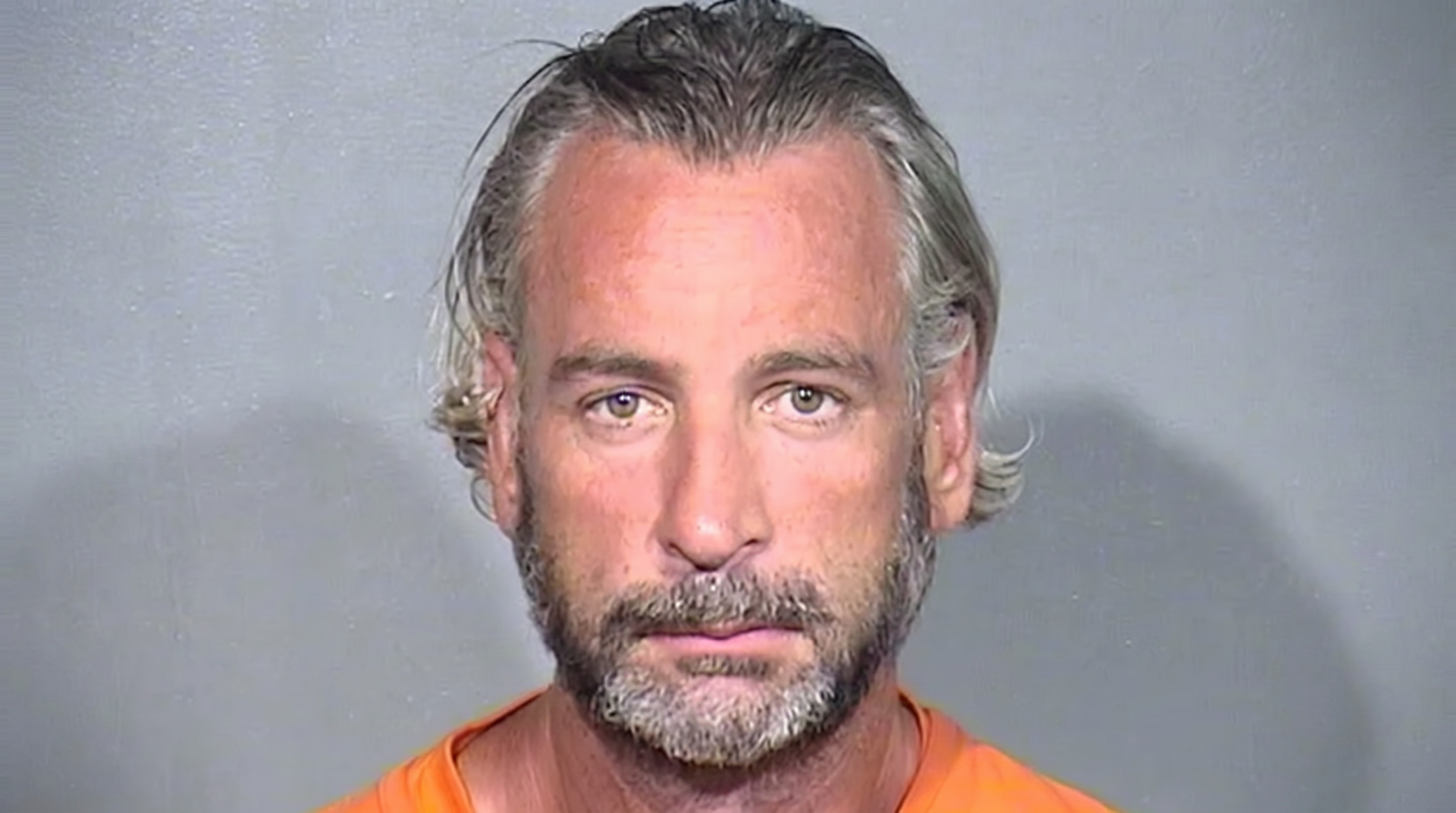 Mugshot of a middle-aged man with gray-blond hair and a salt-and-pepper beard wearing an orange jail jumpsuit.