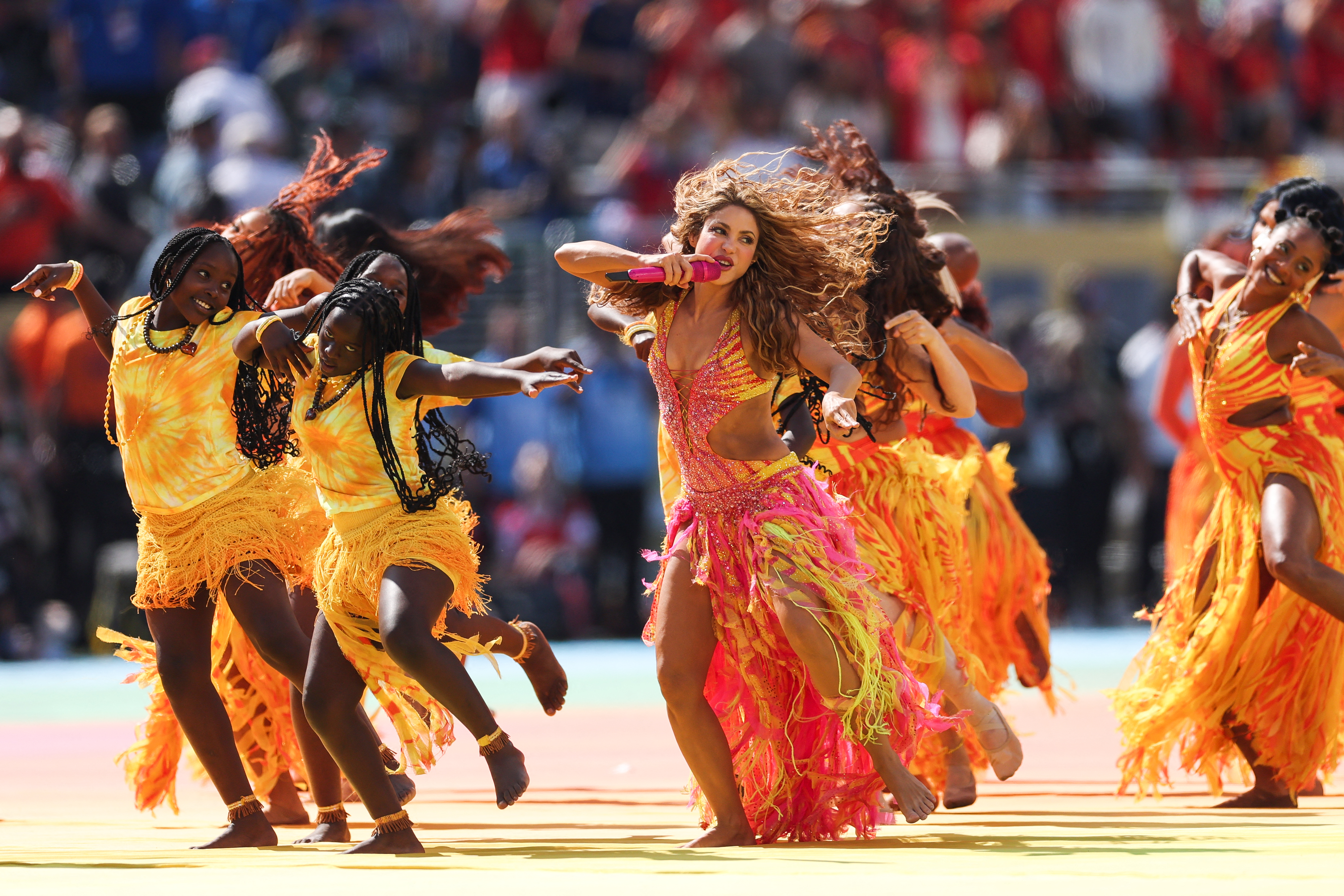 Shakira in a vibrant pink and yellow sequined costume sings into a microphone while dancing energetically with a group of performers in orange fringe outfits at a stadium halftime show.