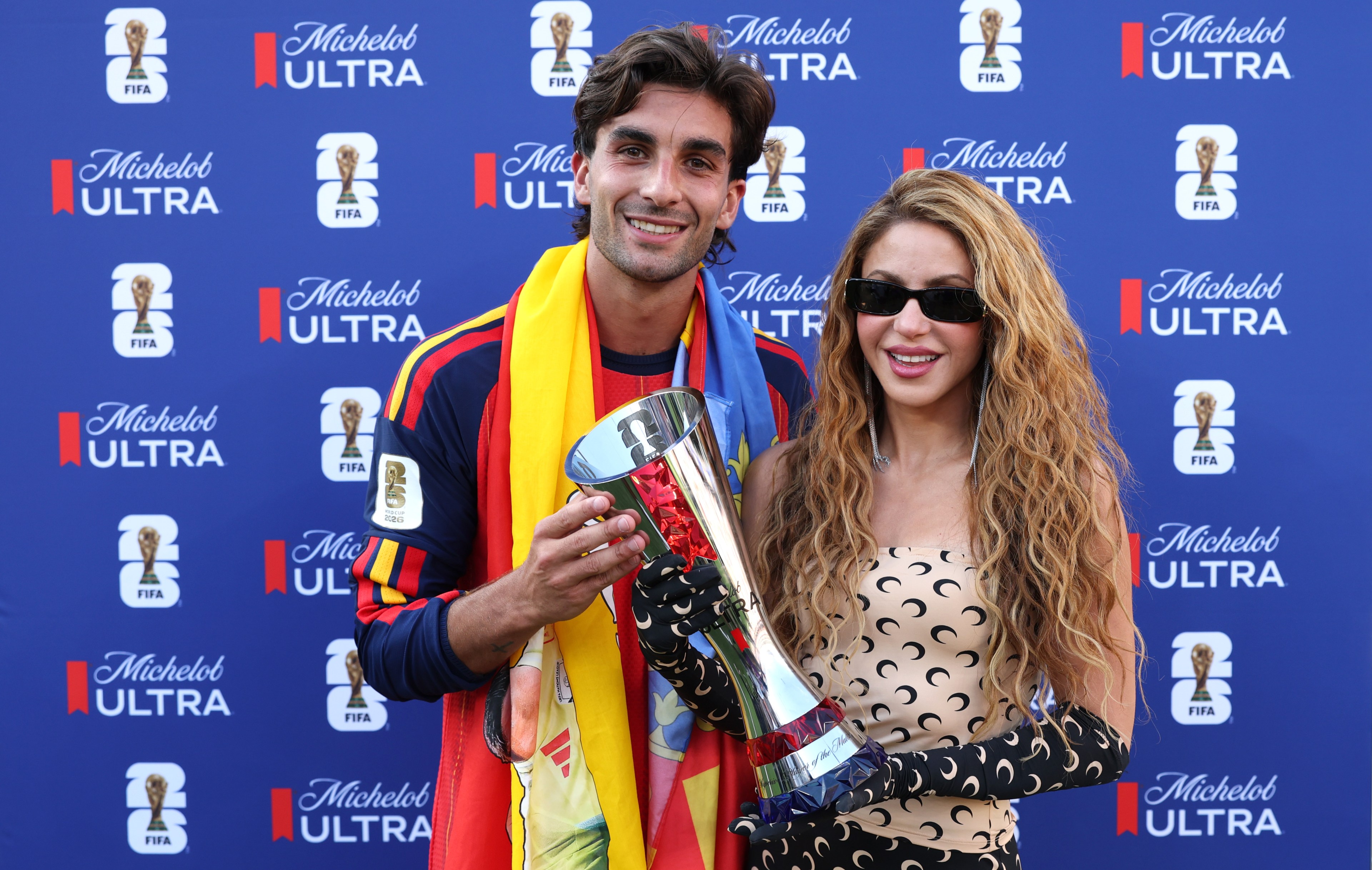 Ferran Torres in a Spanish flag scarf holds a silver trophy next to a woman in sunglasses and a moon-print outfit, posing in front of a FIFA and Michelob Ultra backdrop.