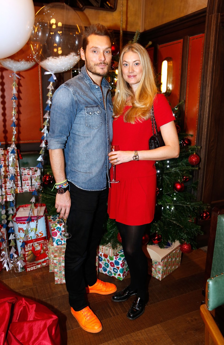 A man in a denim shirt and orange sneakers stands beside a blonde woman in a red dress holding a champagne flute, in front of a decorated Christmas tree.