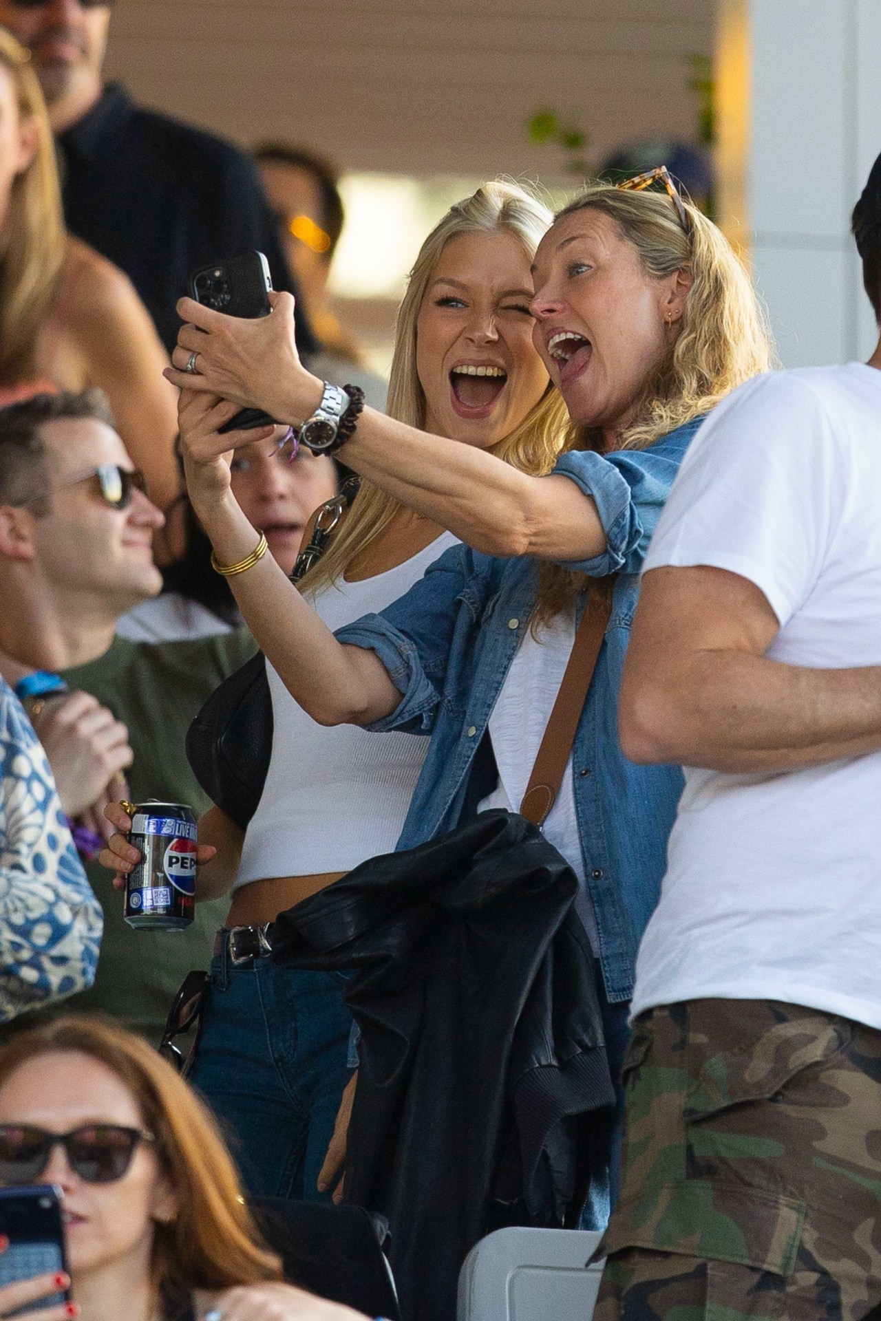Two blonde women laughing and taking a selfie together in a crowd at an outdoor concert.