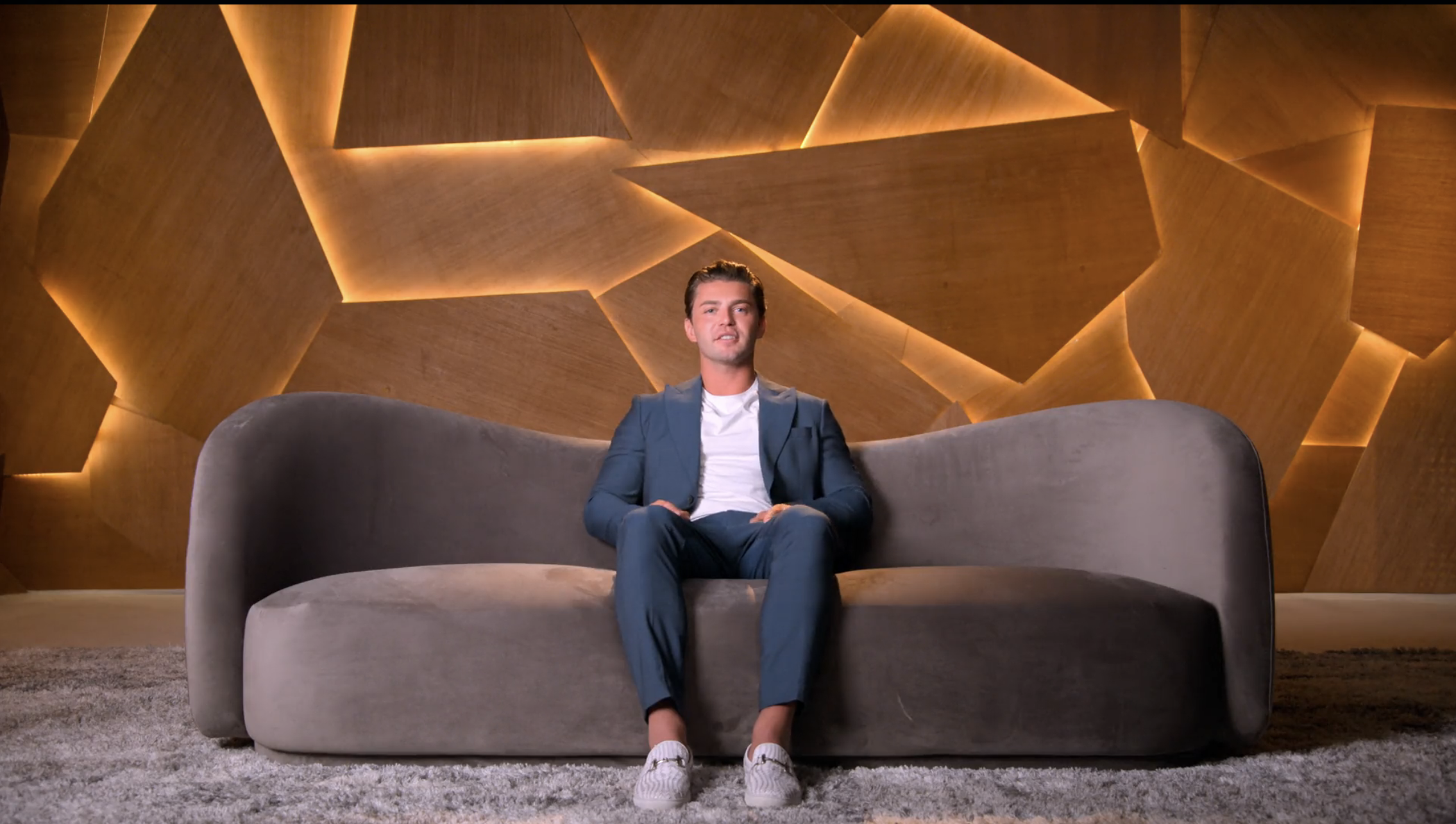 Man in a blue suit and white T-shirt sitting alone on a gray curved sofa in front of a glowing geometric wood-panel wall.