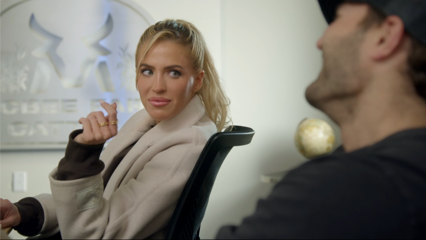 Blonde woman in a beige coat sitting in a black office chair, looking sideways at a man whose back is to the camera, in an office setting.