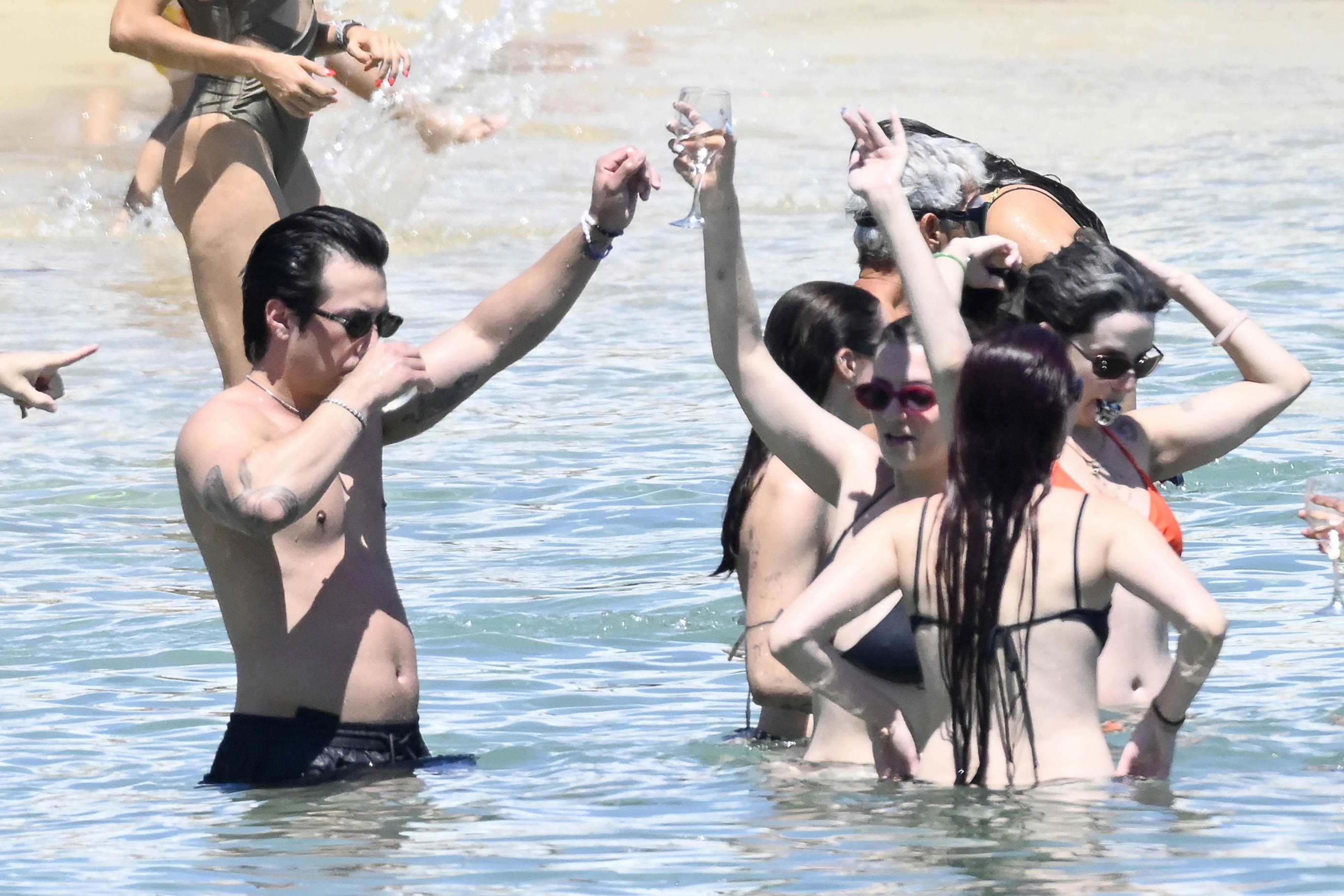 A shirtless tattooed man with dark hair and sunglasses holds a champagne glass aloft in shallow ocean water, surrounded by women in bikinis with arms raised.