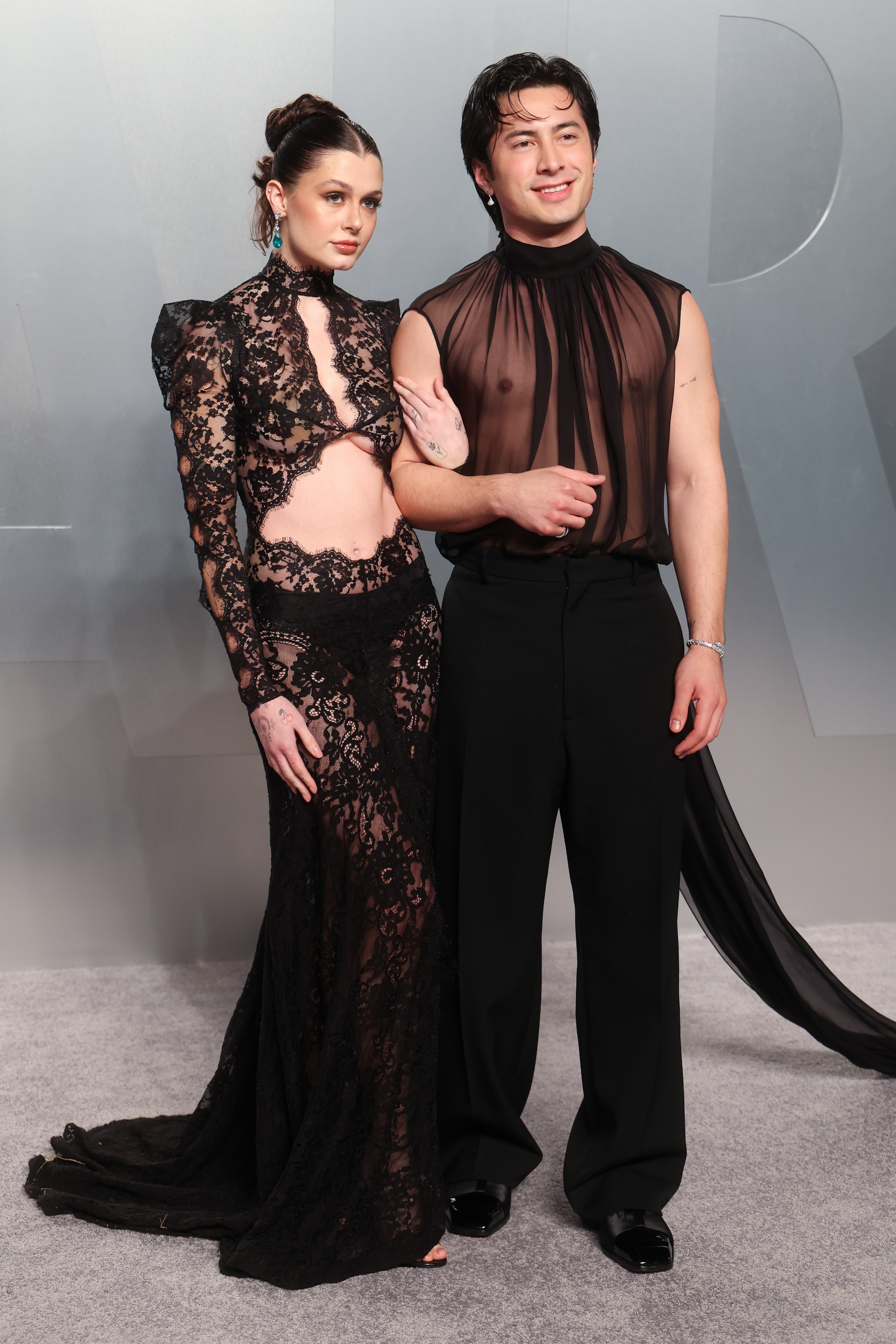 A woman in a black sheer lace gown and a man in a sheer black sleeveless top with black trousers pose together on a gray carpet.