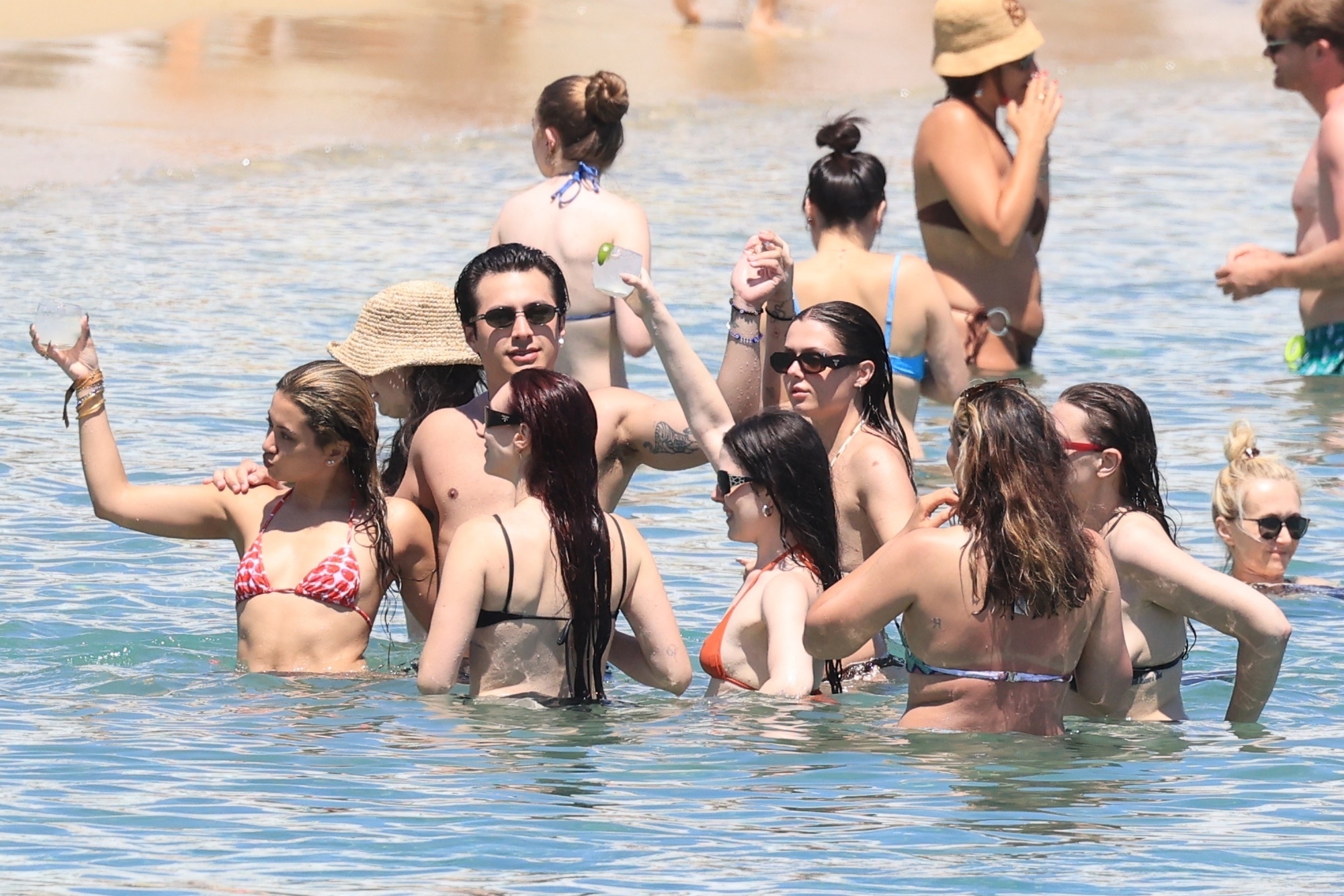 A group of people in bikinis and swim trunks gathered waist-deep in clear ocean water at a beach, with one man in sunglasses and a straw-hatted woman among them.