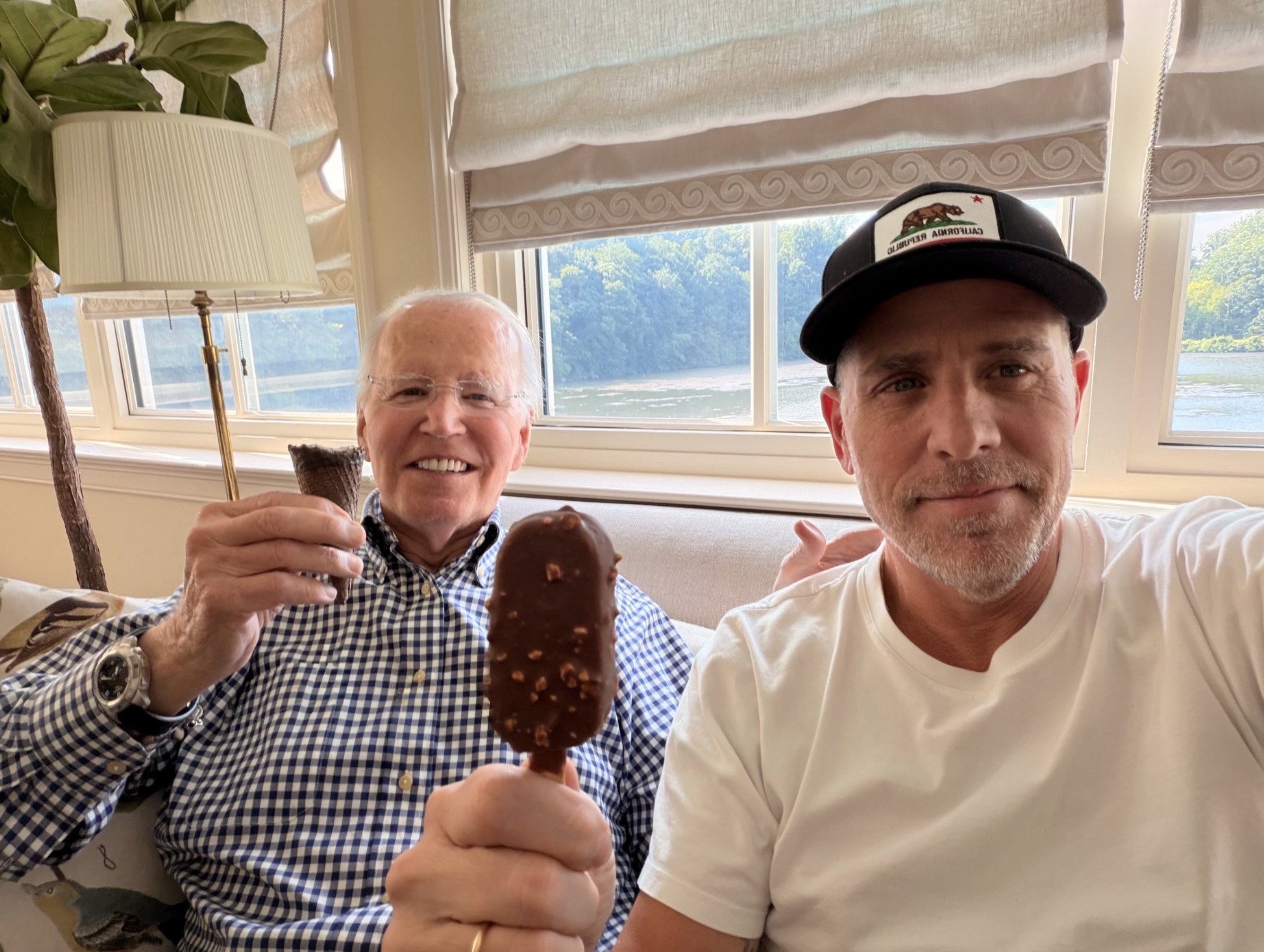 Two men sitting on a couch smiling, one holding up a chocolate-covered ice cream bar on a stick, the other holding a cone, with a California Republic cap on one man.