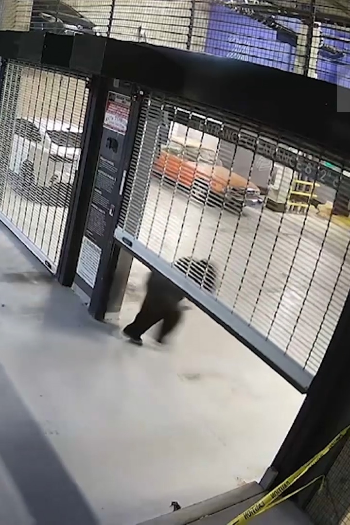 Grainy security camera footage of a person in dark clothing walking through a gated apartment complex entrance at night.