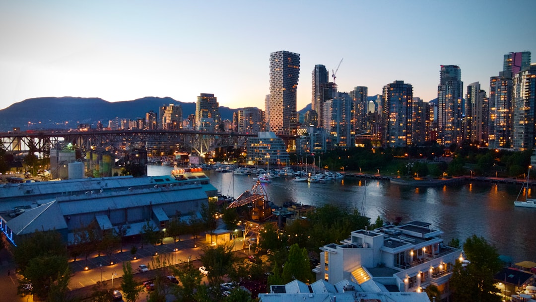 Aerial dusk view of Vancouver's False Creek, with marina boats, Granville Bridge, and illuminated downtown skyscrapers backed by mountains.