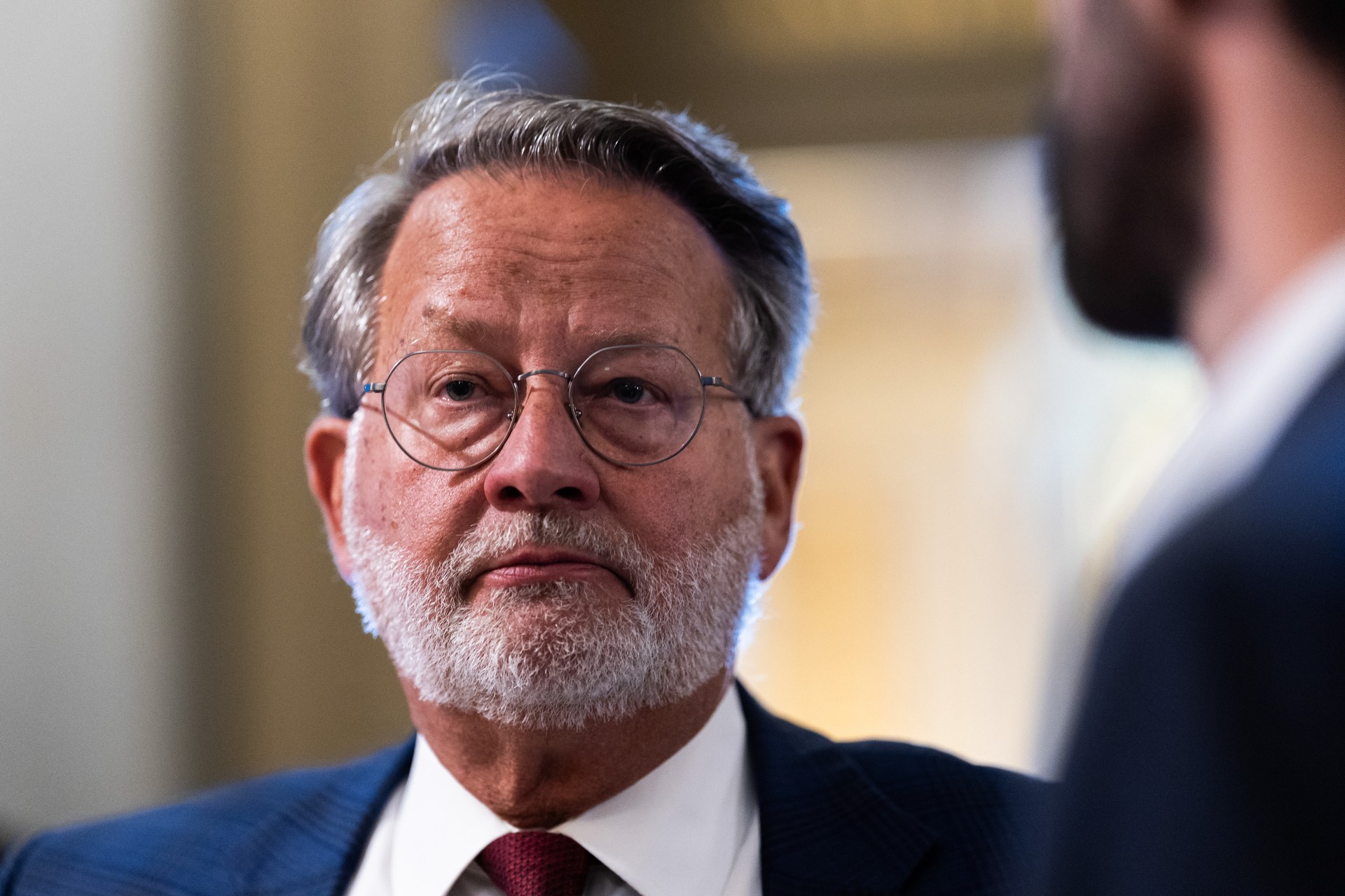 Sen. Gary Peters wearing round glasses, a navy suit, and a red tie, looking serious inside the U.S. Capitol Building.