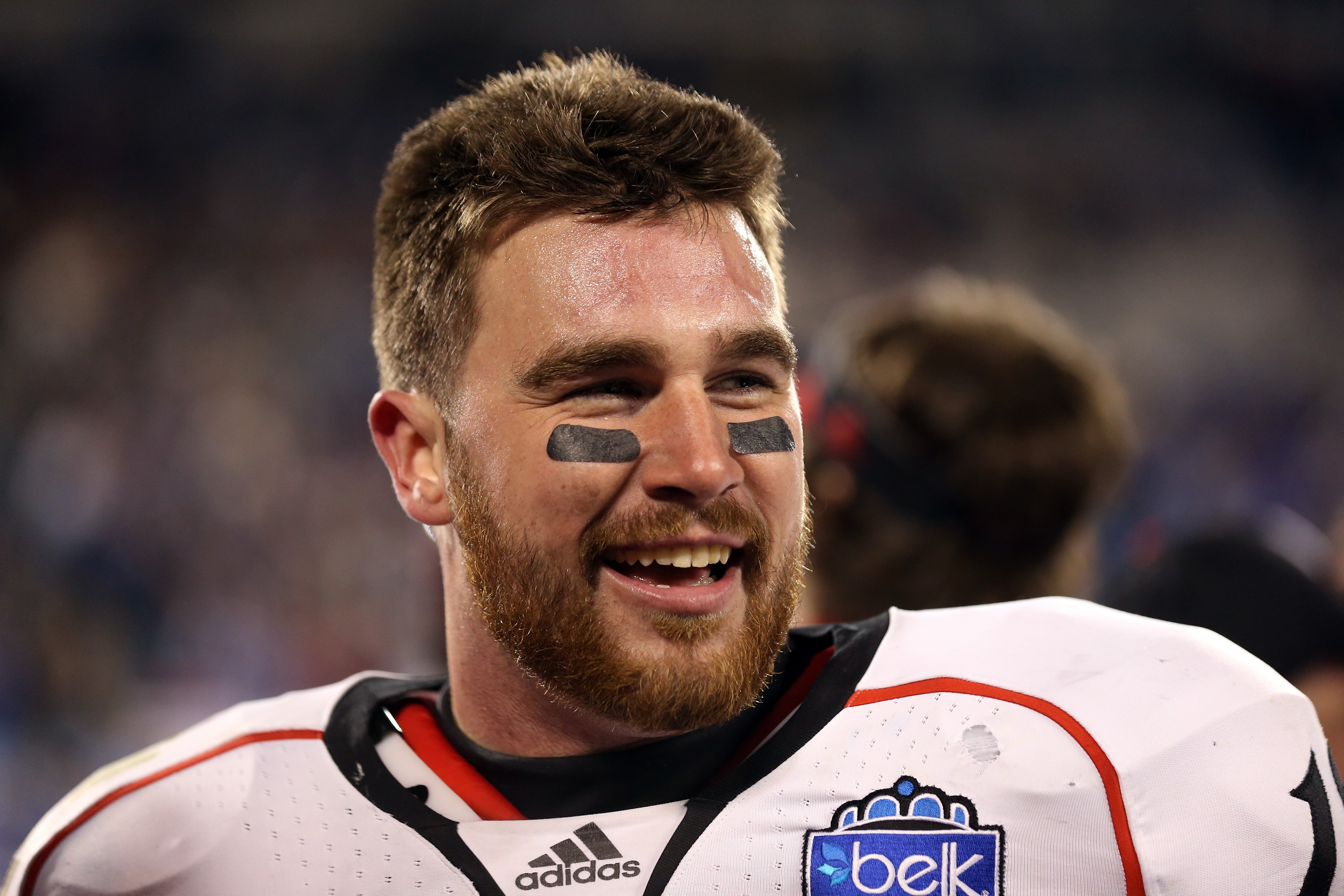 Football player in white Cincinnati Bearcats uniform with eye black smiling on the sideline after a game.