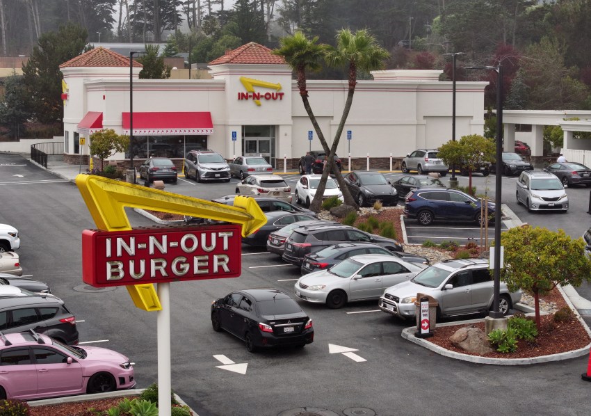 Aerial view of an In-N-Out Burger restaurant in Daly City, California, with a full parking lot and the red-and-yellow In-N-Out Burger sign in the foreground.