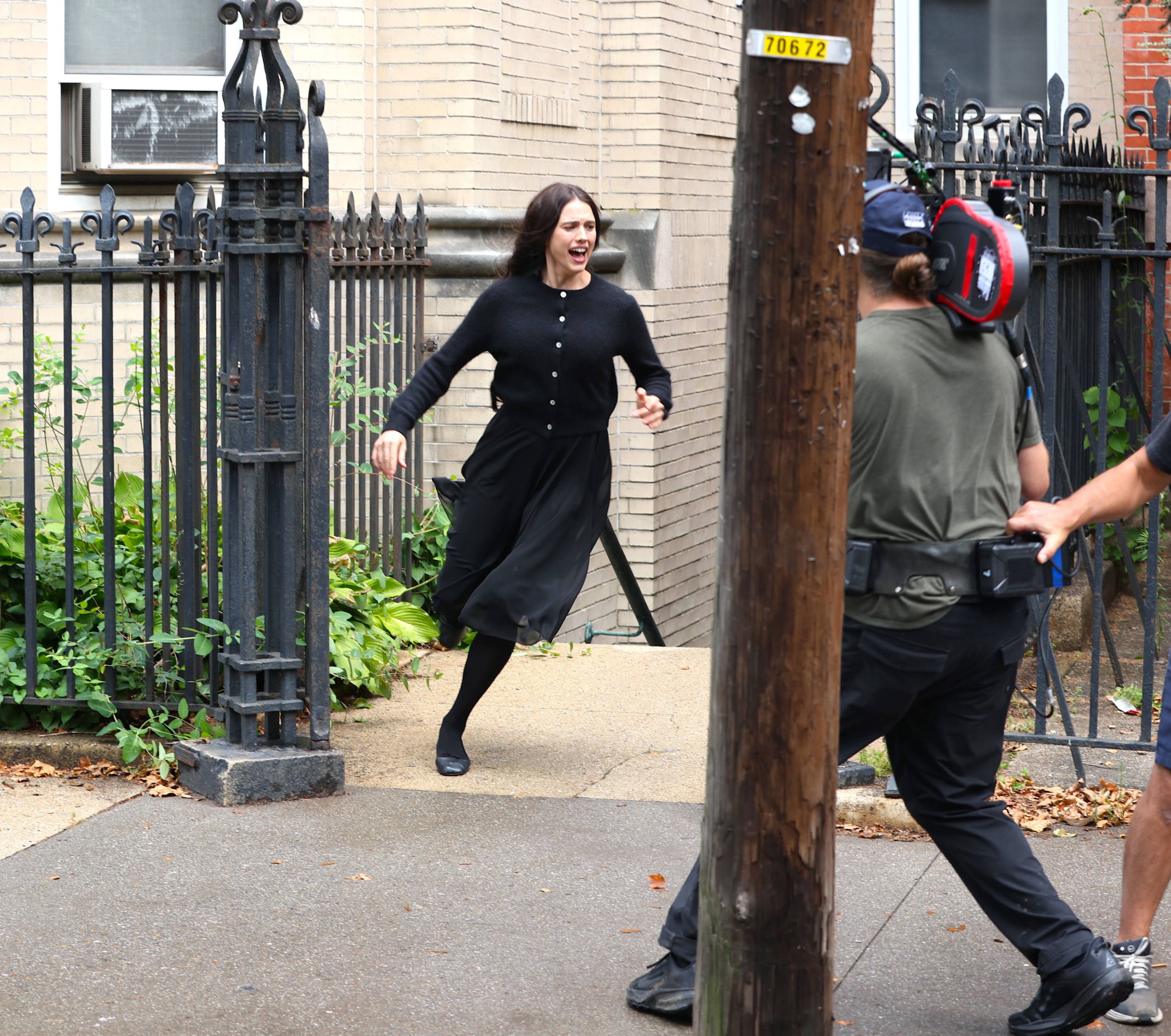 Actress in black dress and cardigan runs and screams on a city sidewalk near an iron fence while a camera operator films her.