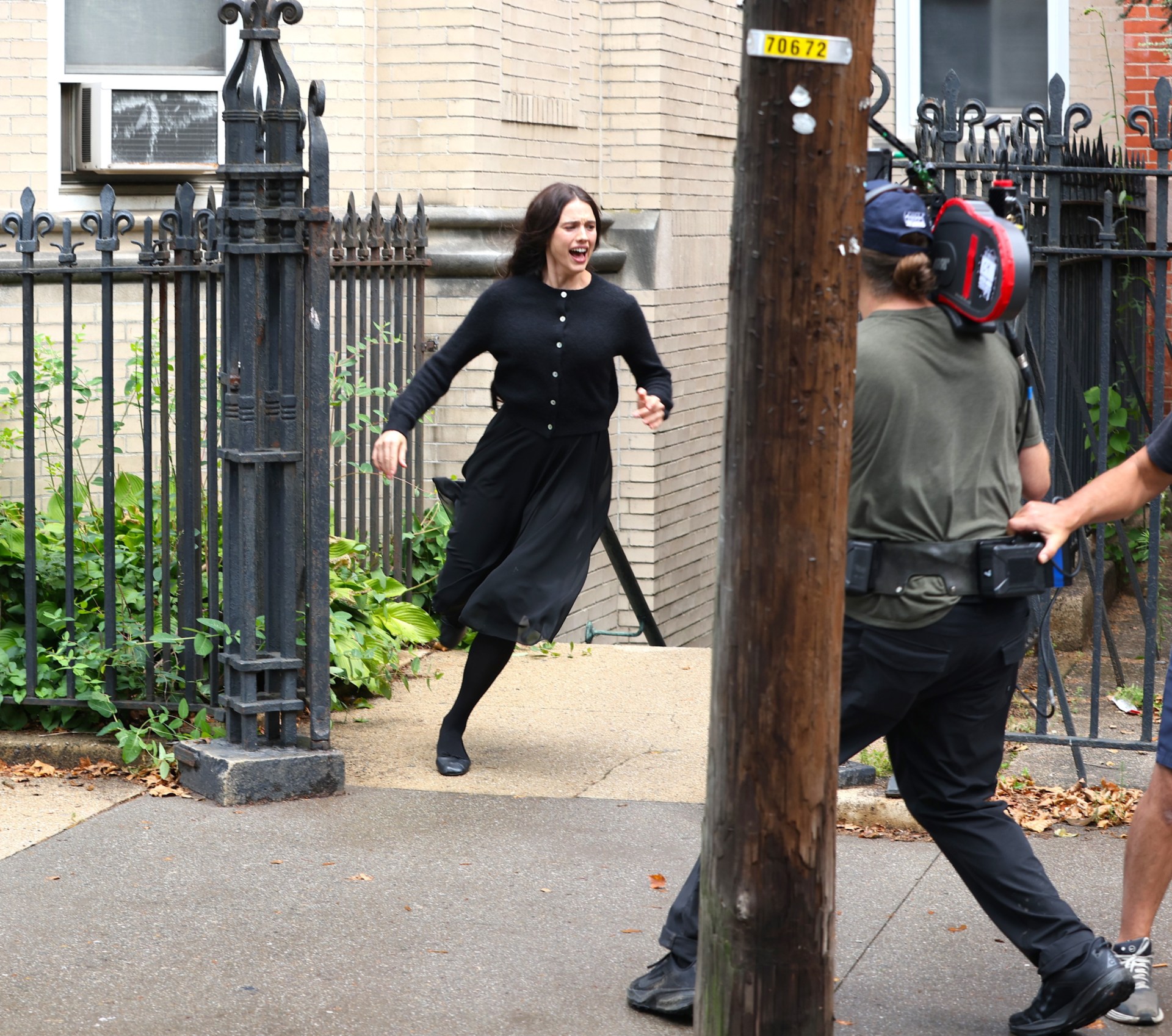 Actress in black dress and cardigan runs and screams on a city sidewalk near an iron fence while a camera operator films her.
