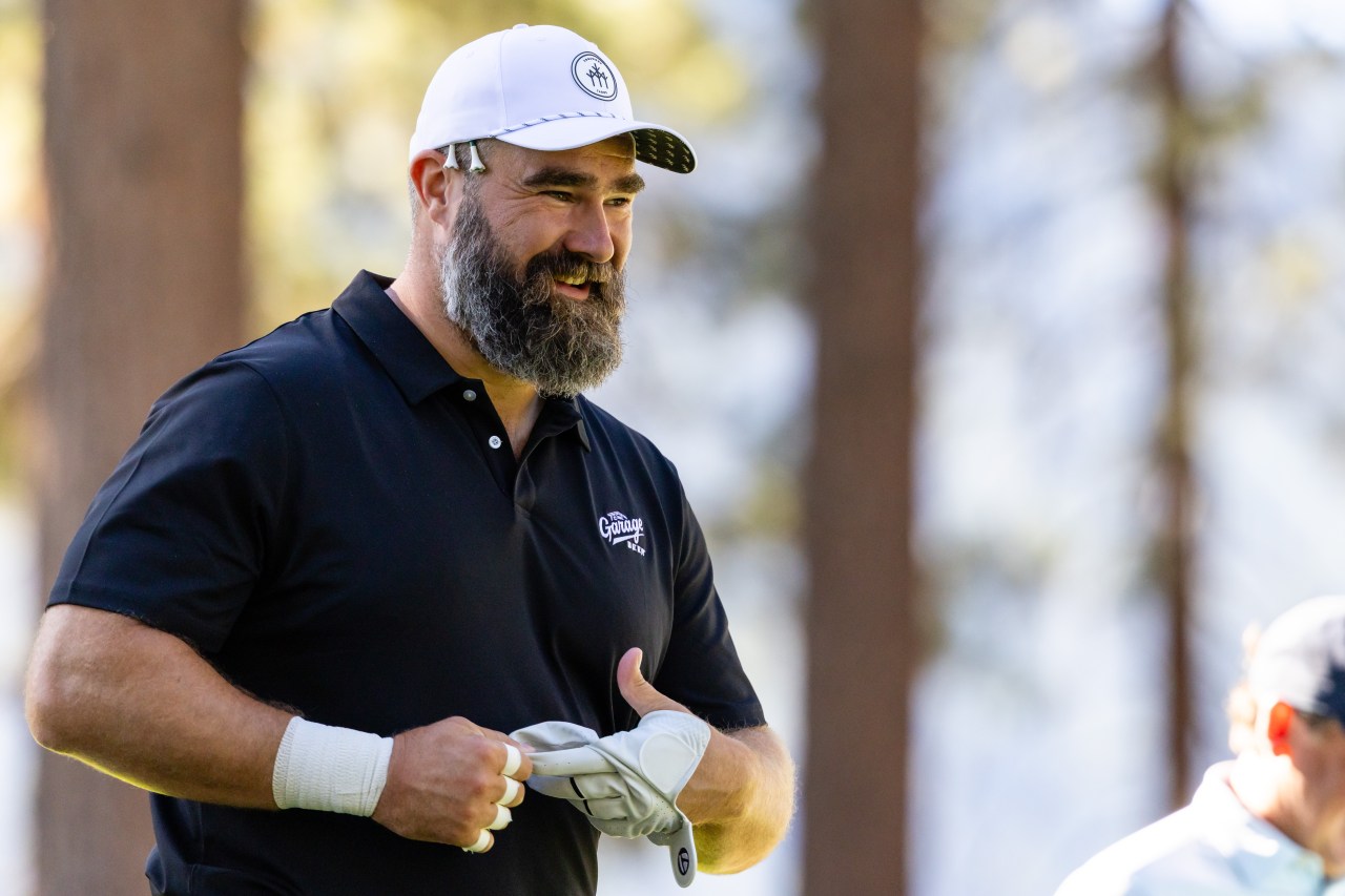 Bearded man in a white cap and black polo shirt smiling and adjusting a golf glove on a golf course.