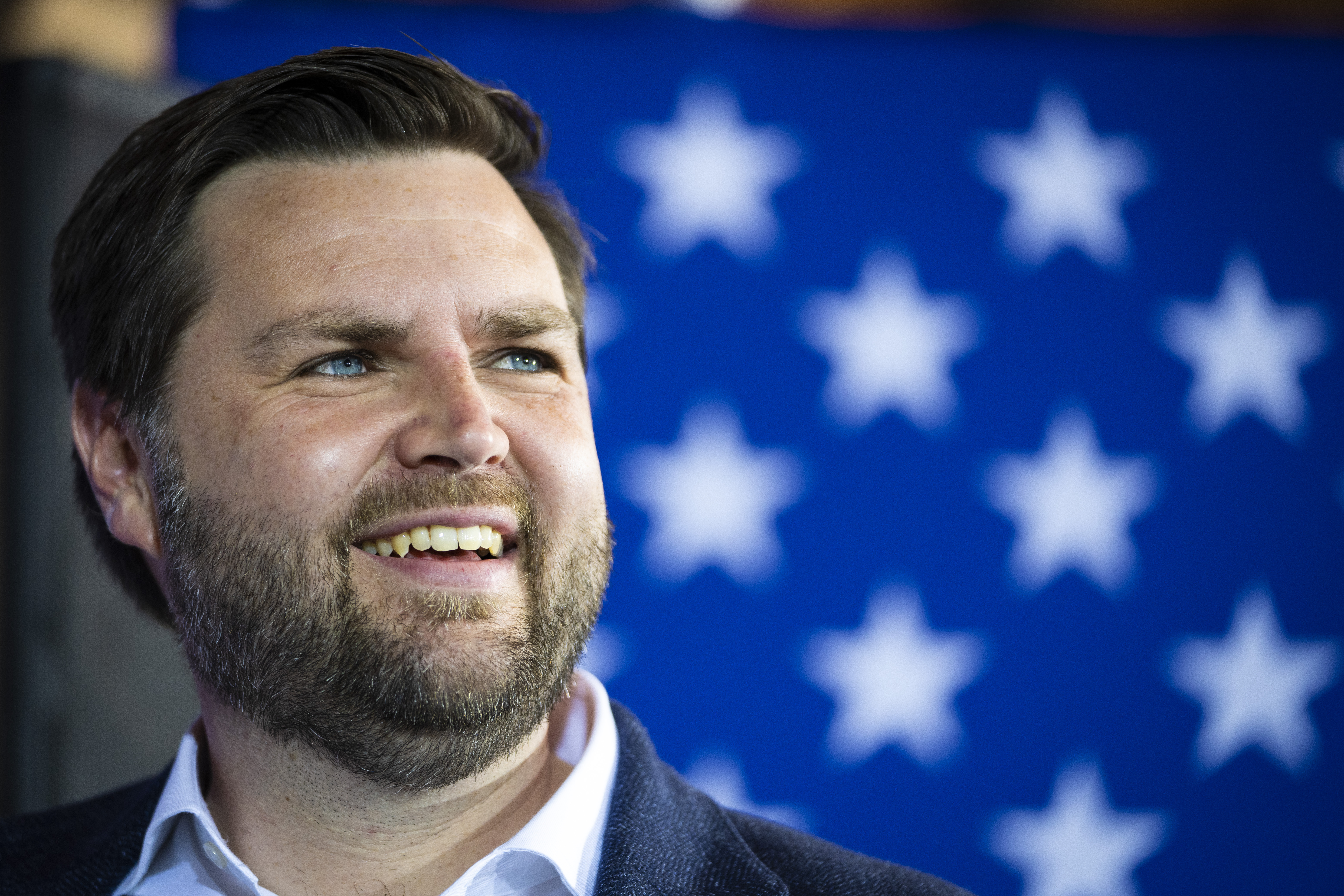 Close-up of a smiling man with dark hair and a beard in front of a blue background with white stars.