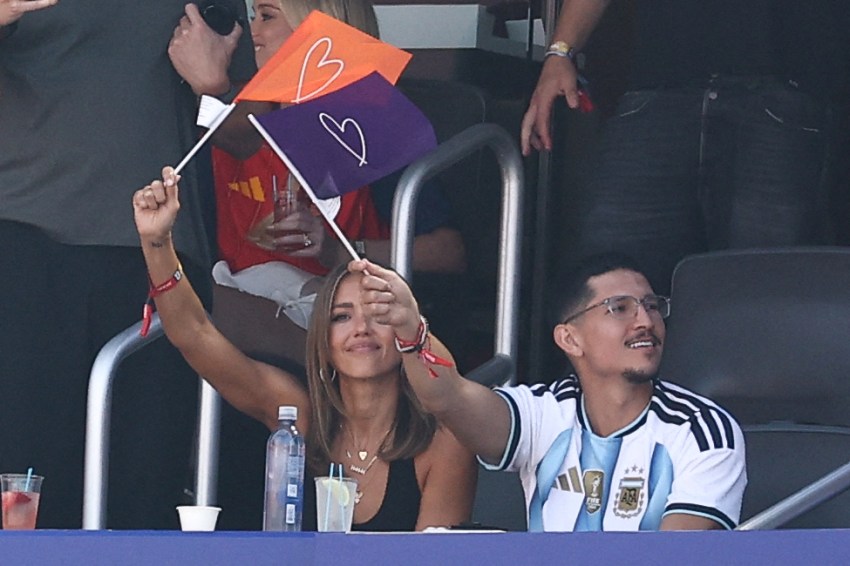 A woman waves two small flags with heart designs while a man in an Argentina jersey sits beside her in stadium stands.