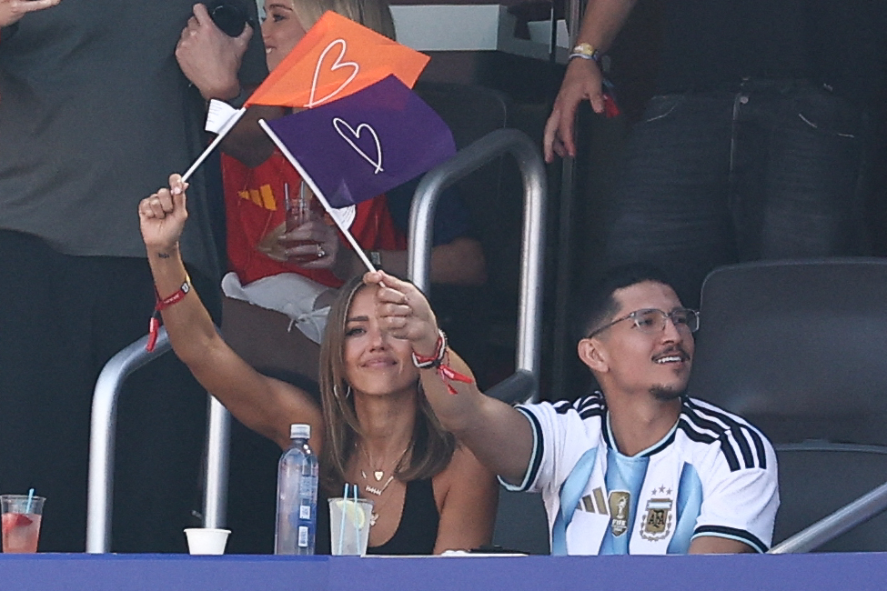A woman waves two small flags with heart designs while a man in an Argentina jersey sits beside her in stadium stands.
