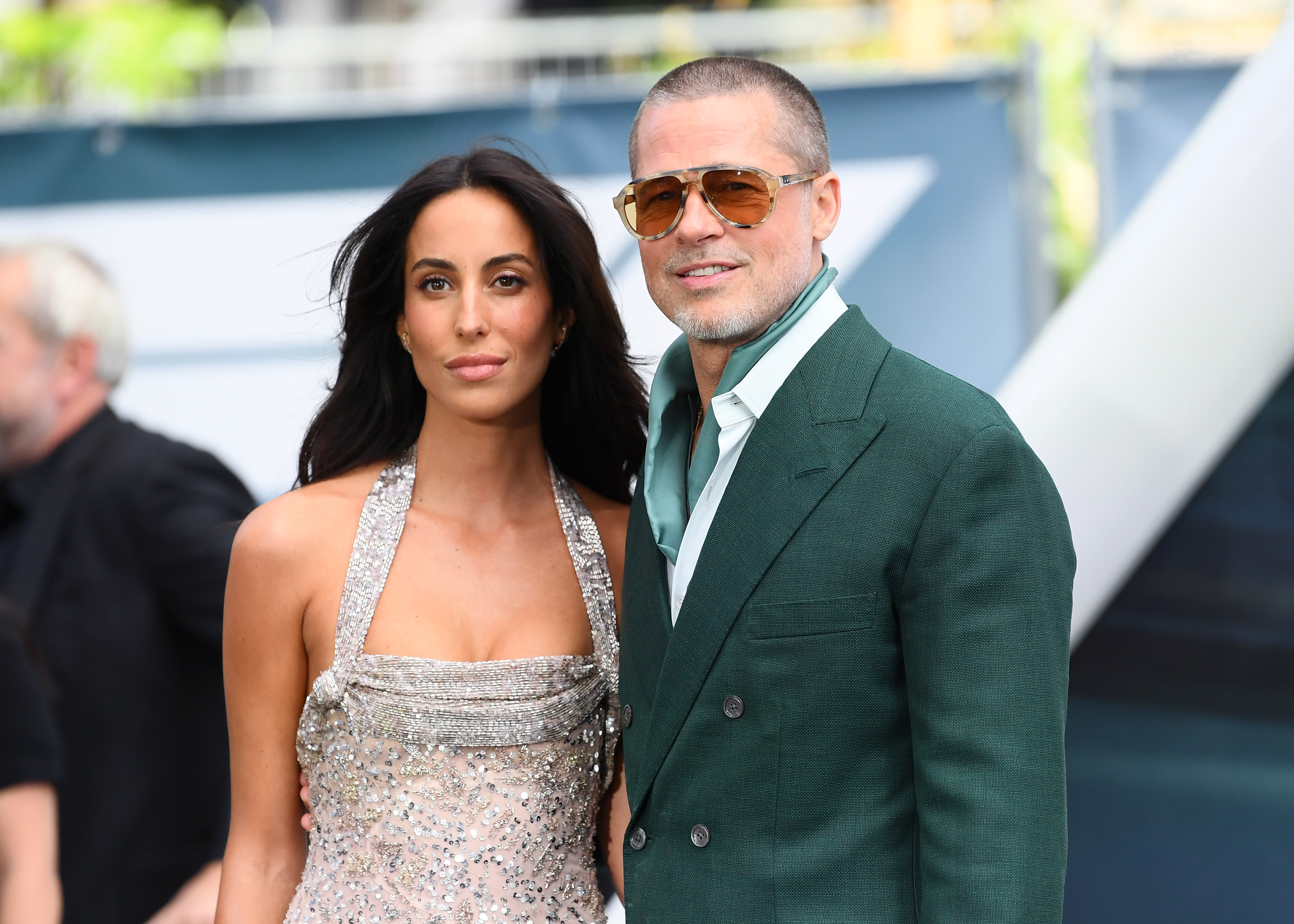 Ines de Ramon in a silver sequined halter gown stands beside Brad Pitt in a green double-breasted suit and tinted aviator sunglasses at a movie premiere.