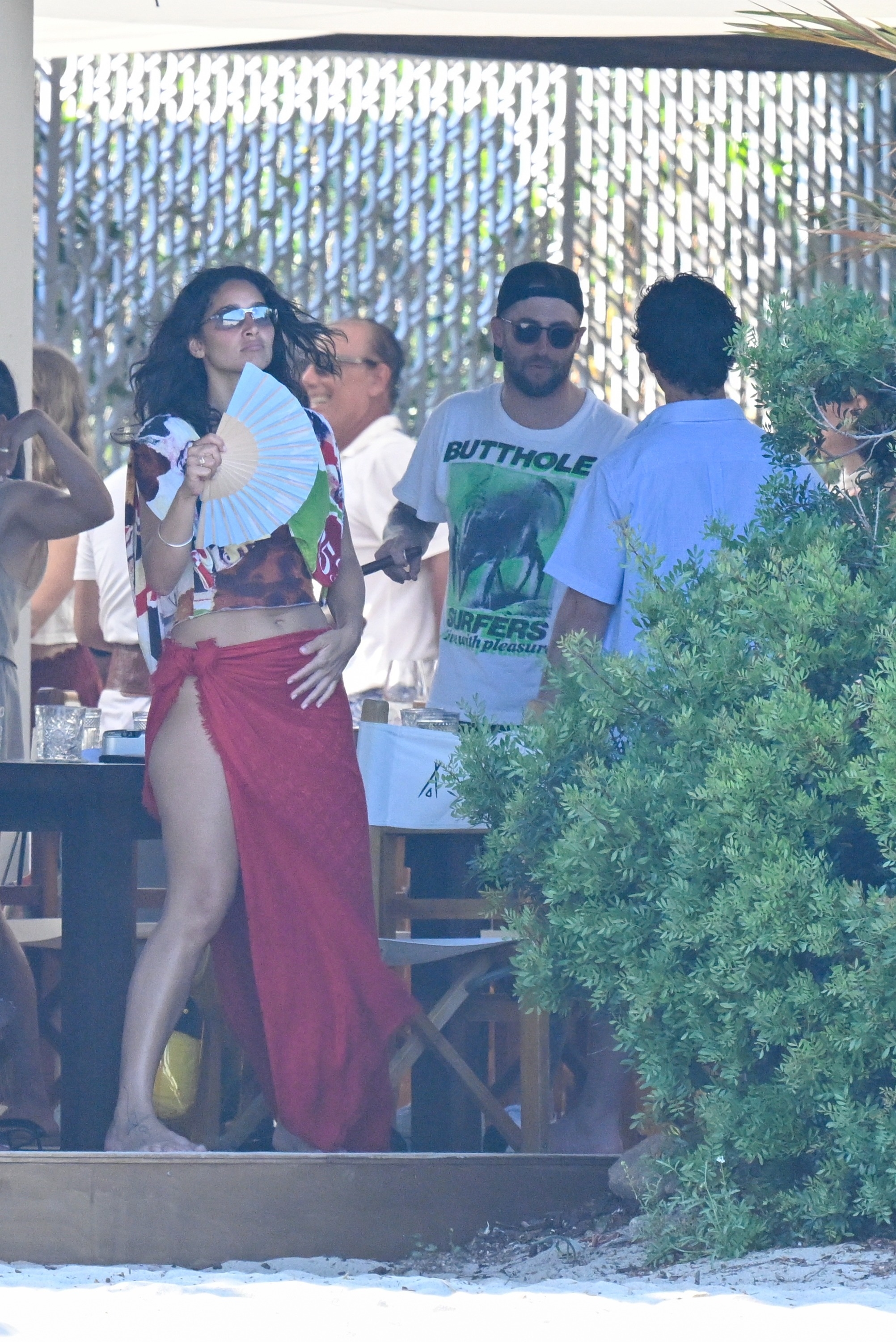 Woman in red wrap skirt holding a paper fan stands beside a man in a white graphic tee at a crowded outdoor beachside venue.
