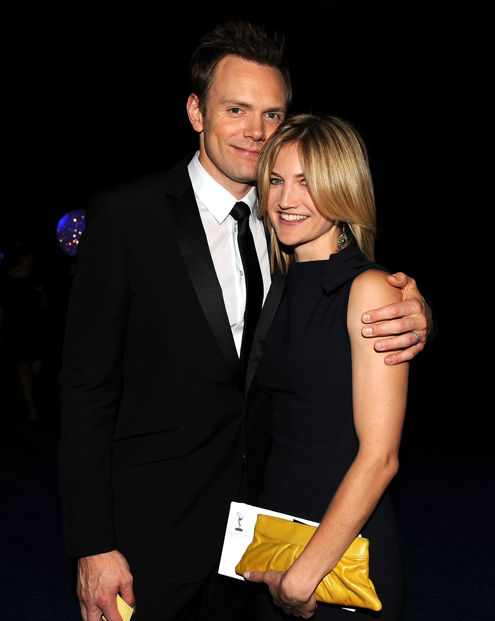 Joel McHale in a black tuxedo with his arm around Sarah Williams in a black dress holding a yellow clutch at the Emmy Awards Governors Ball.