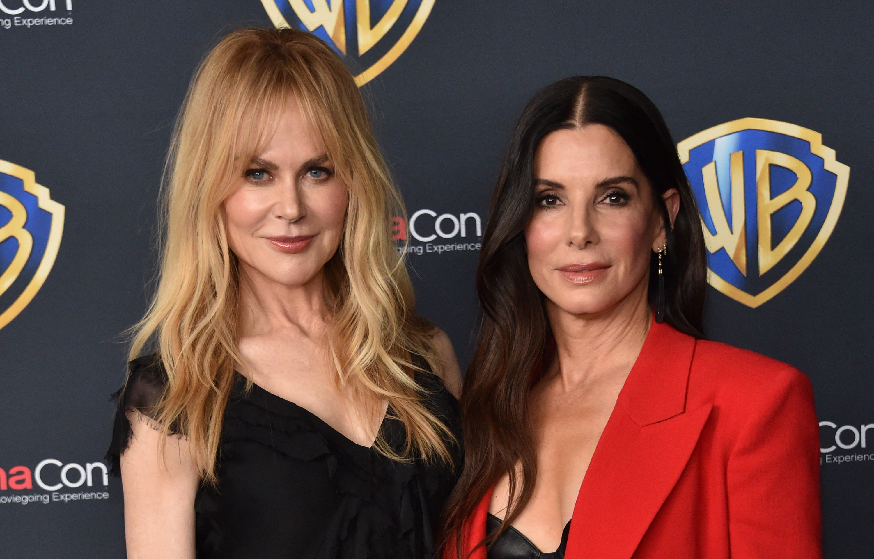 Two women stand in front of a Warner Bros. logo backdrop; one in a black dress with long blonde hair, the other in a red blazer with dark hair.